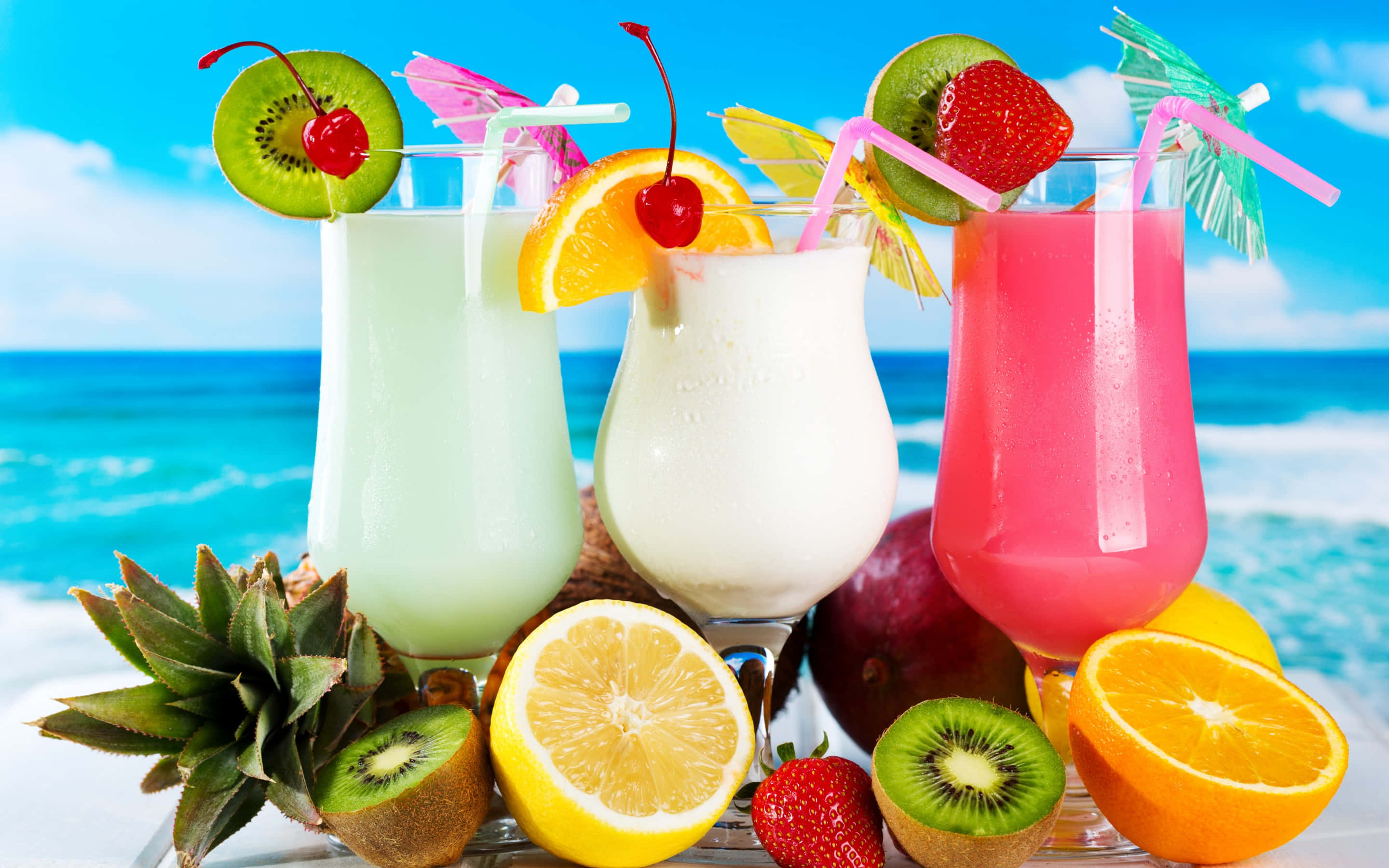 Food And Beverage 桌布 壁紙 — 三杯熱帶水果飲品，飾以奇異果、櫻桃和橙，放在沙灘桌上，旁邊有切片柑橘和鳳梨，背景是明亮的藍色海洋。