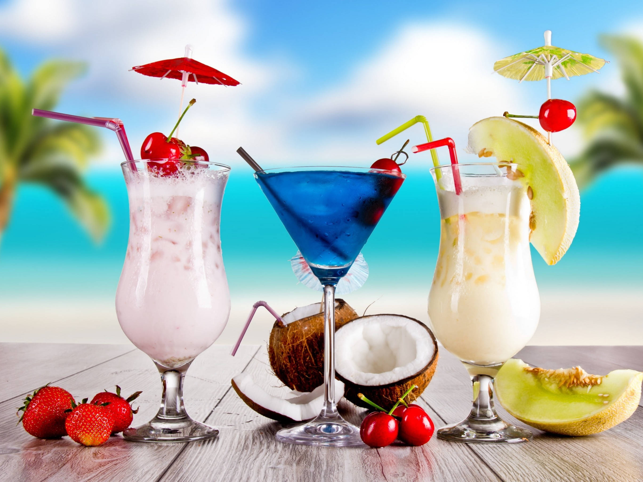 Tropical Drink 桌布 壁紙 — 三杯熱帶雞尾酒，配有傘、櫻桃和水果裝飾，放在木桌上，背景是模糊的海灘。