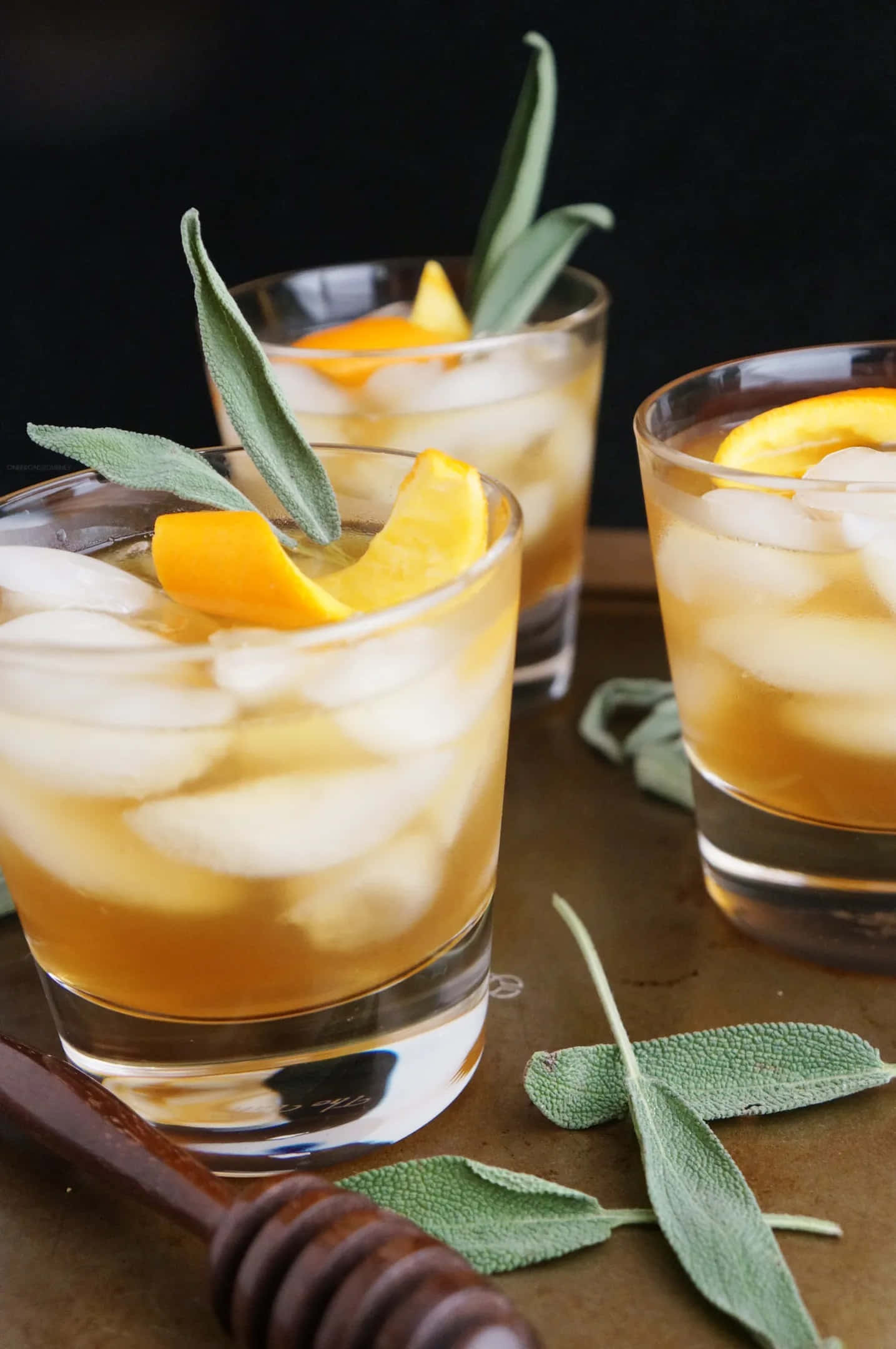 Winter Drinks 桌布 壁紙 — 三杯琥珀色雞尾酒配上冰塊、橙片和鼠尾草葉，放在一個質樸的托盤上，旁邊有一個木製蜂蜜棒。