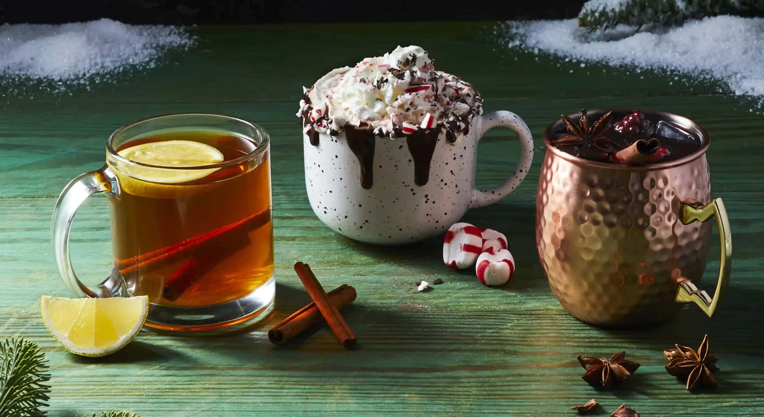 Winter Drinks 桌布 壁紙 — 三杯節日冬季飲品，配上鮮奶油、糖果手杖和香料，放在一個白雪覆蓋的綠色木桌上。