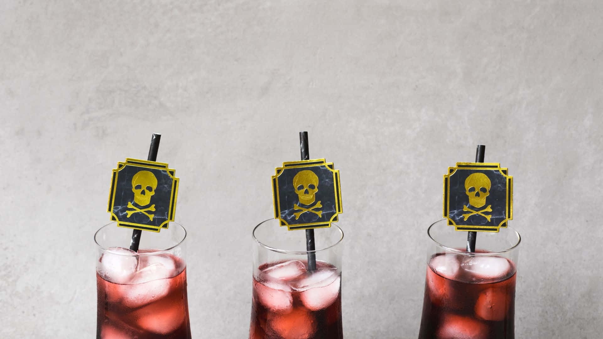 Spooky Cocktails 桌布 壁紙 — 三杯紅色冰飲，配有骷髏頭和交叉骨攪拌棒，背景為有紋理的灰色。