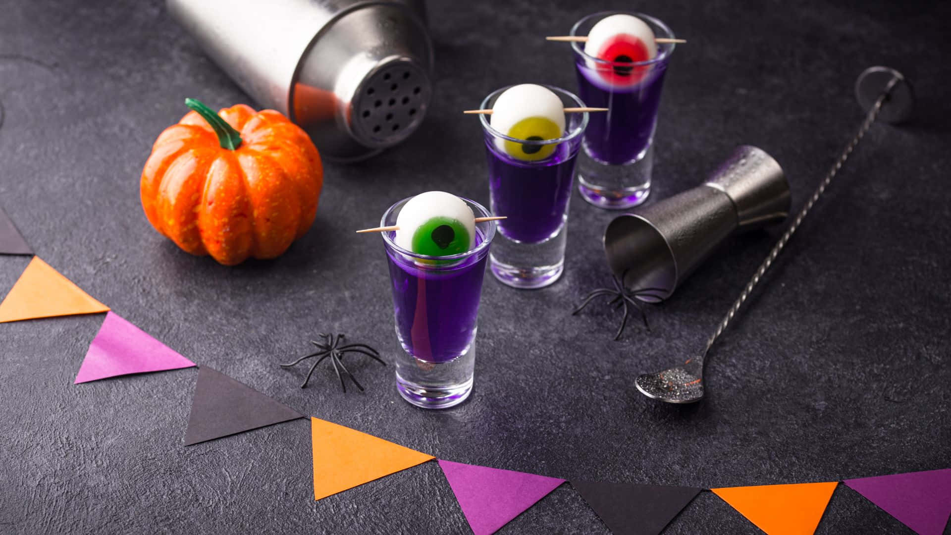Spooky Cocktails 桌布 壁紙 — 三杯紫色萬聖節主題的 shots，上面放有糖果眼睛，旁邊有一個南瓜和雞尾酒工具，放在深色表面上。