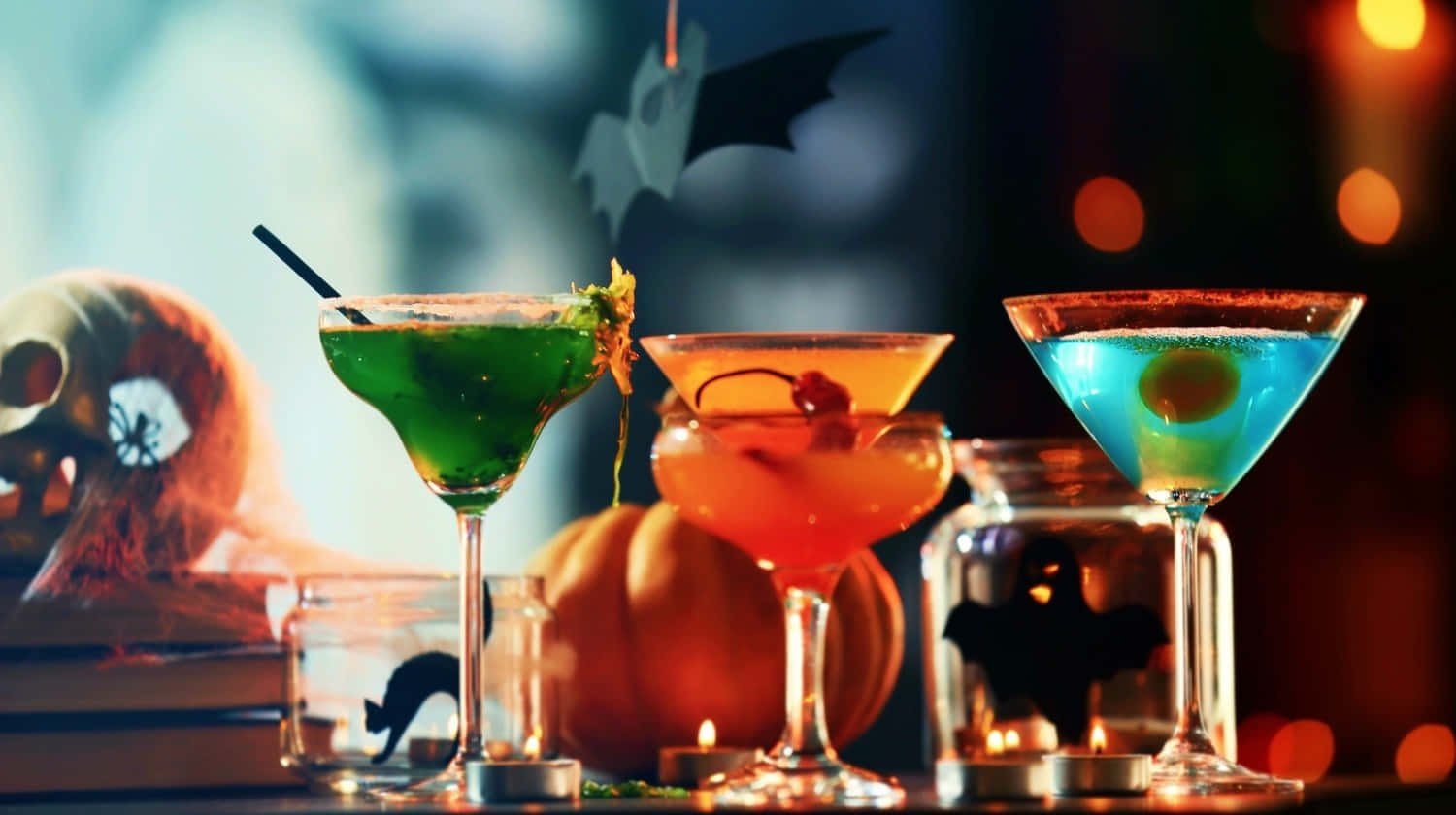 Spooky Cocktails 桌布 壁紙 — 三杯色彩繽紛的萬聖節雞尾酒盛載於馬丁尼杯中，放在桌上，旁邊有南瓜和鬼怪裝飾，在溫暖的燭光下。