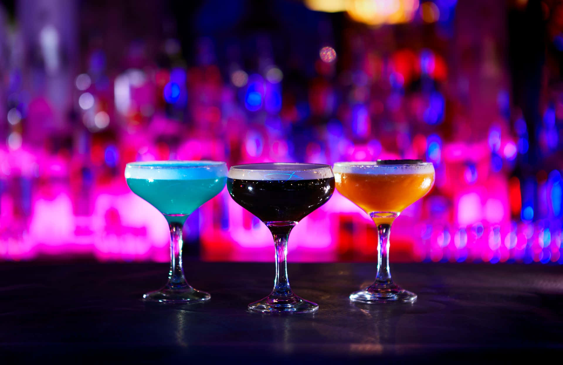 drinks 桌布 壁紙 — 三杯色彩繽紛的雞尾酒盛載於庫佩杯中，在黑暗酒吧檯上的霓虹燈下閃耀。