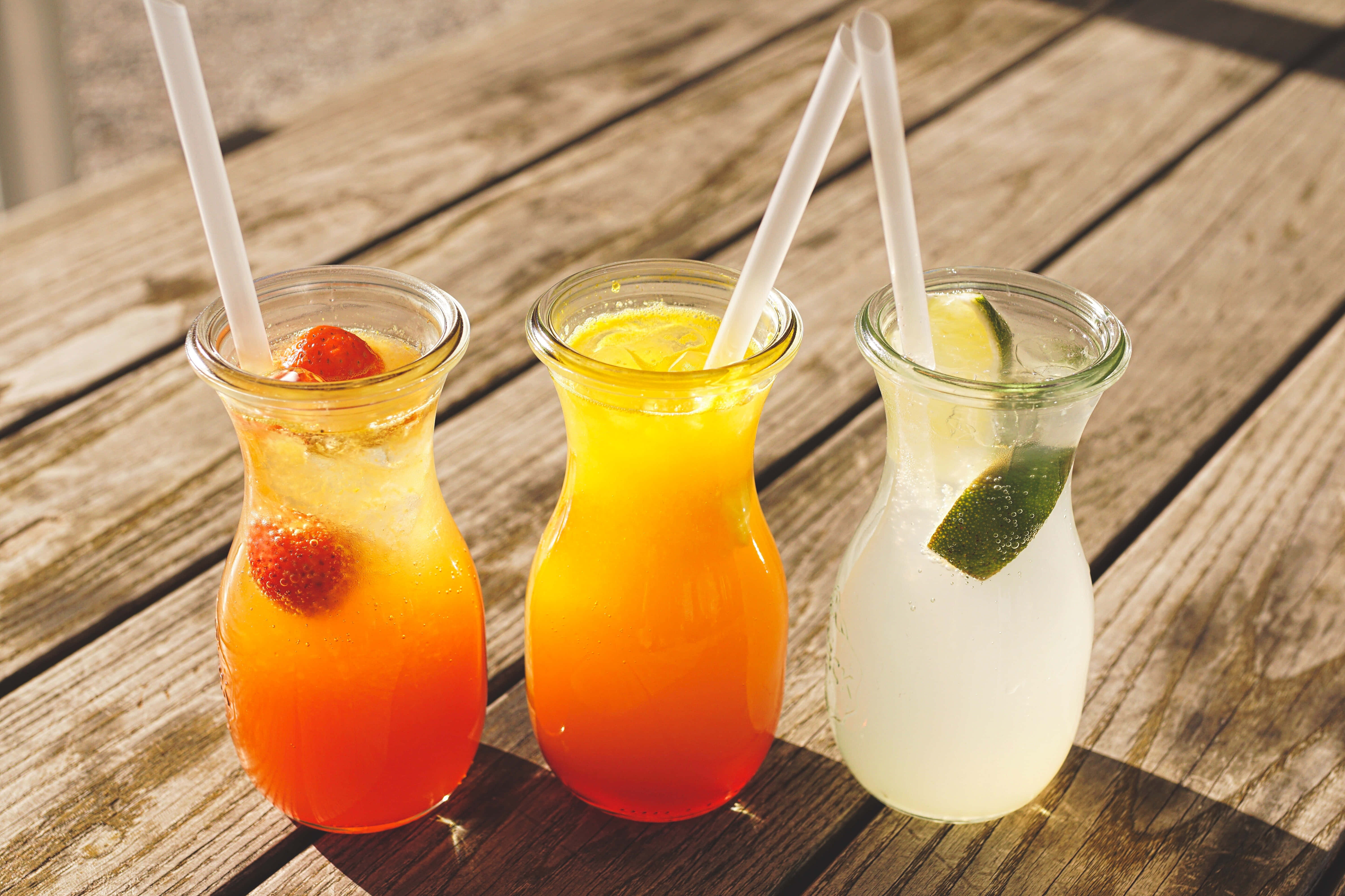 Summer Drinks 桌布 壁紙 — 三杯色彩鮮豔的雞尾酒裝在玻璃瓶中，附有吸管和水果裝飾，放在一張經風化的木桌上，在明亮的陽光下。