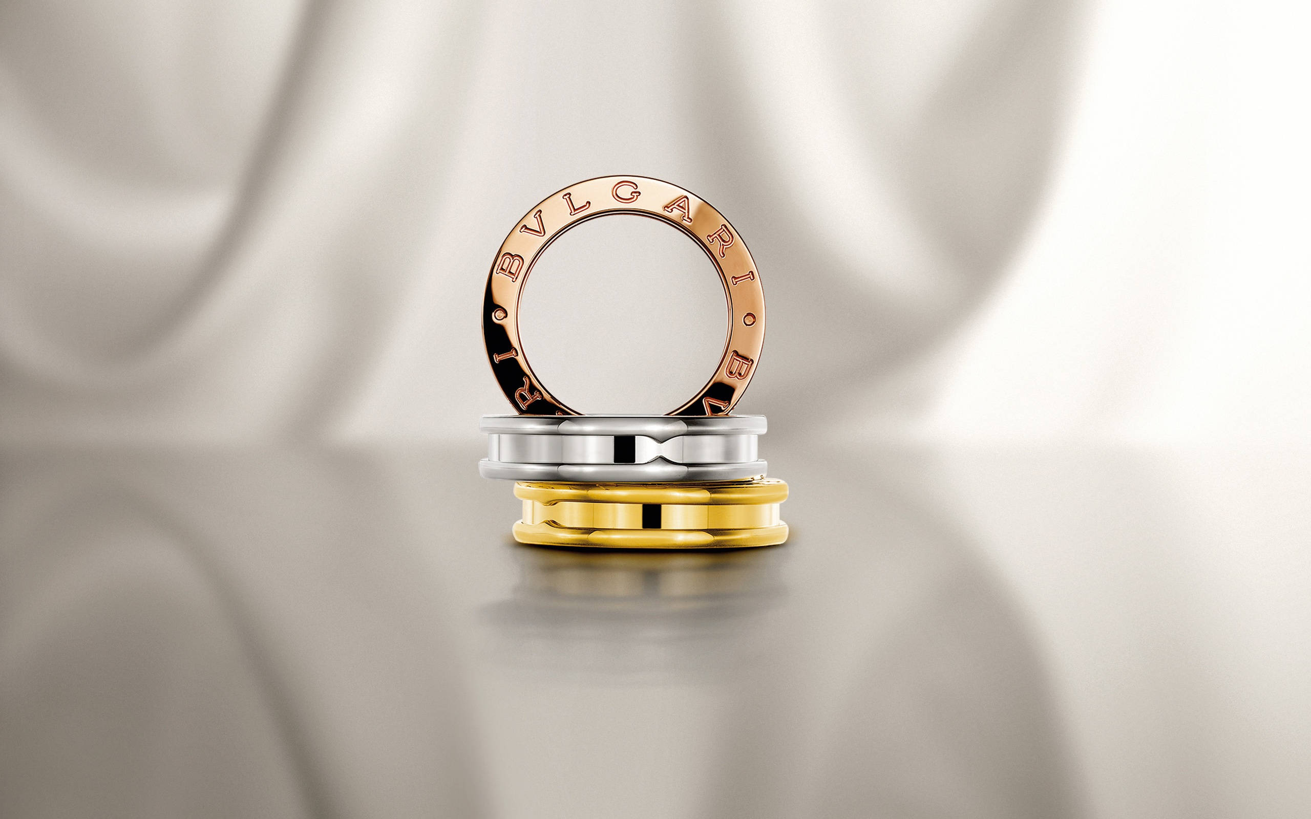 Bvlgari 桌布 壁紙 — 三枚Bvlgari戒指，分別為玫瑰金、銀色和黃金，堆疊在帶有垂褶布料背景的反射表面上。