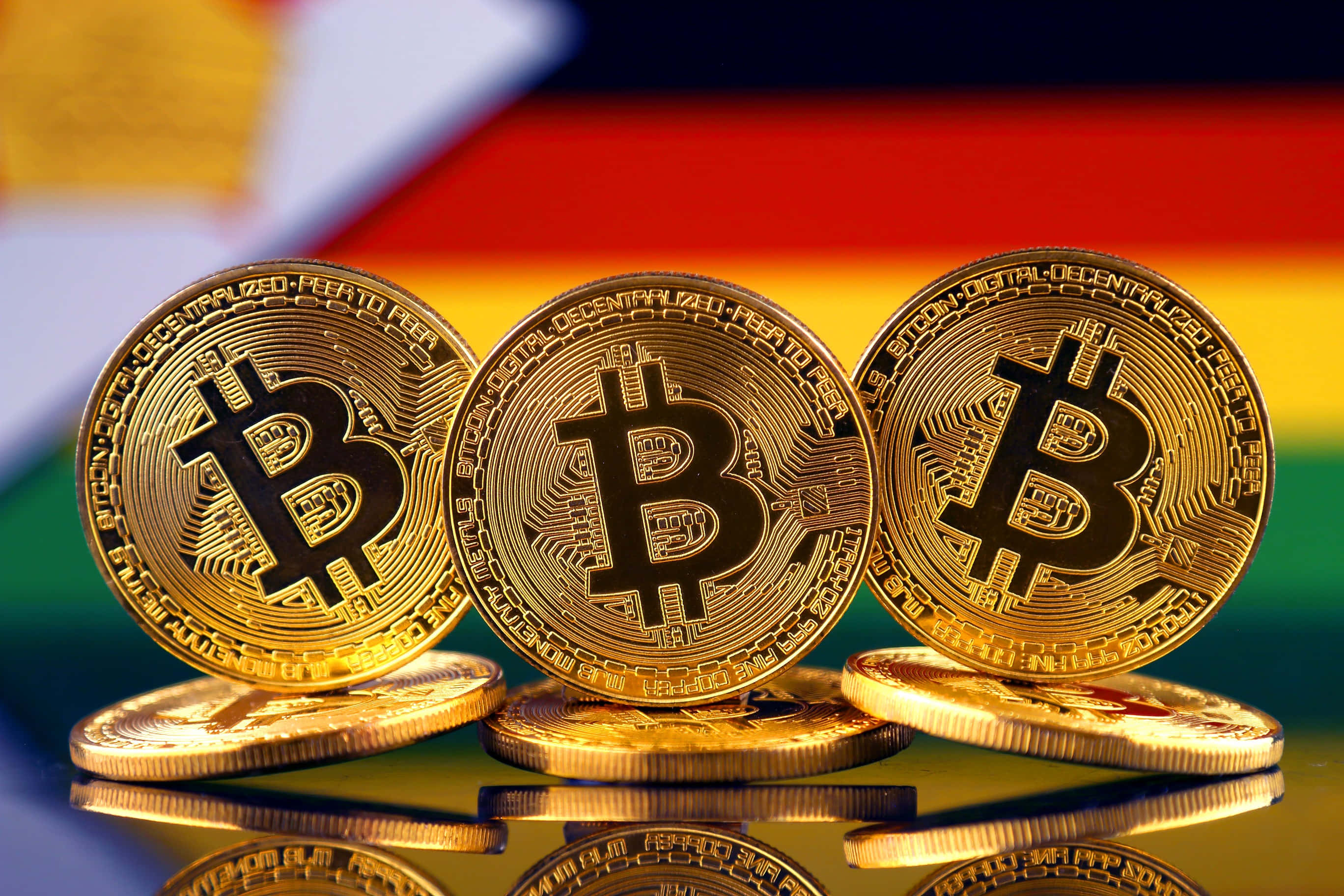 比特幣 桌布 壁紙 — 三枚閃閃發光的金色比特幣硬幣排成一排，放在反光表面上，背景是模糊的德國國旗。