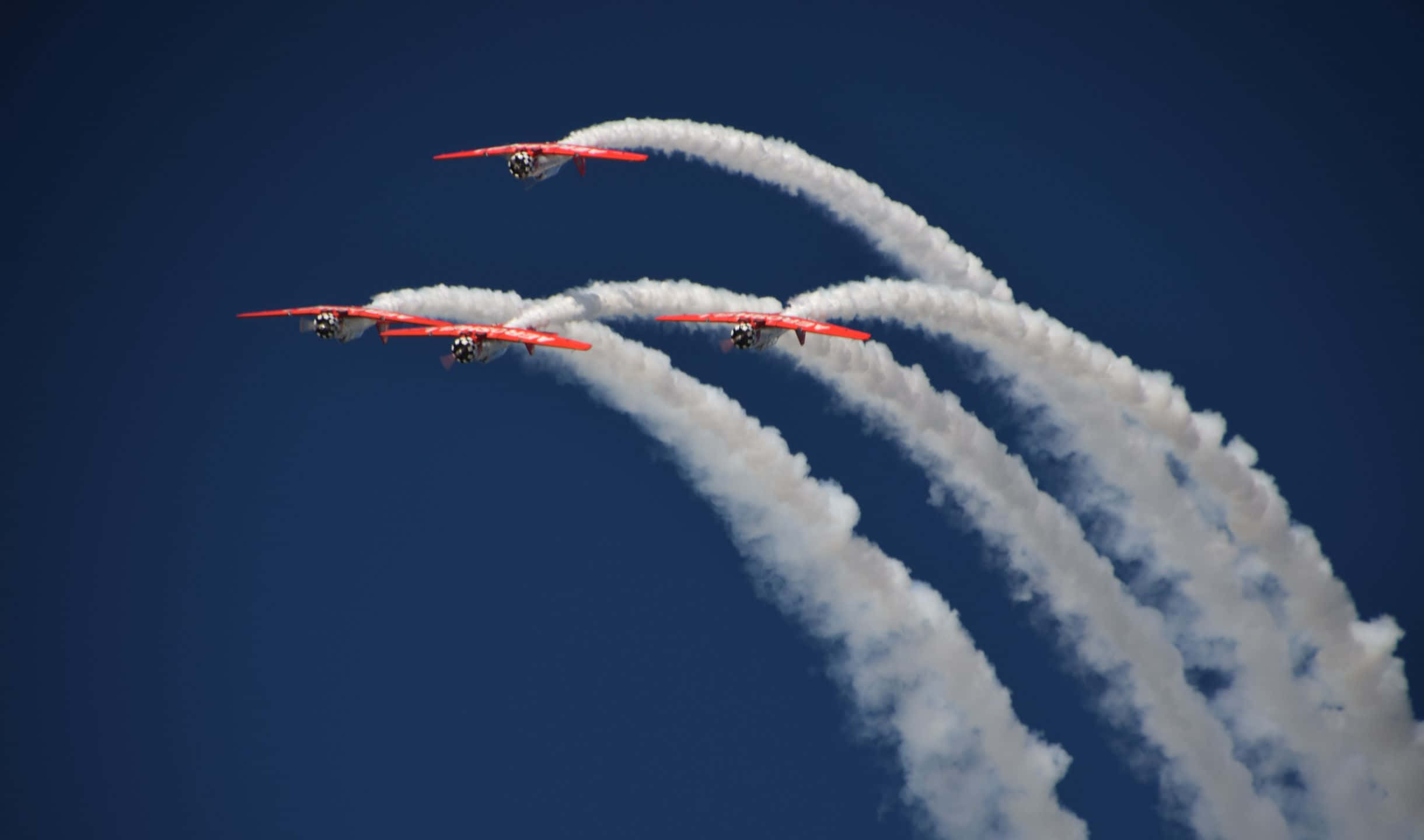 2440x1440 Small Planes 桌布 壁紙 — 三架紅色雙翼機以緊密編隊翱翔於深藍色天空，留下濃厚的白色煙霧軌跡。