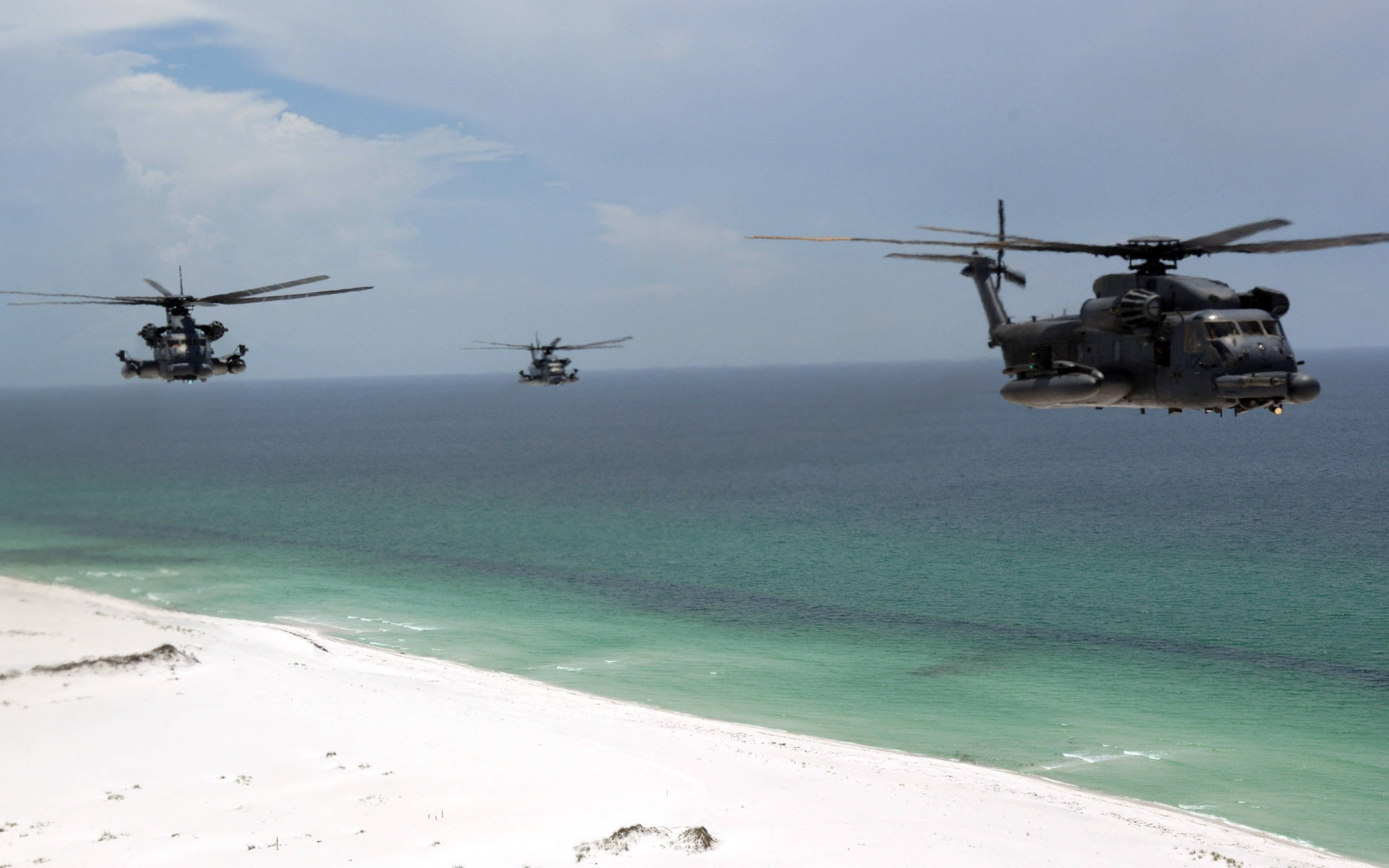 Helicopter 桌布 壁紙 — 三架軍用直升機低飛過一片純淨的白色沙灘，與碧綠色的海洋相接，天空部分多雲。