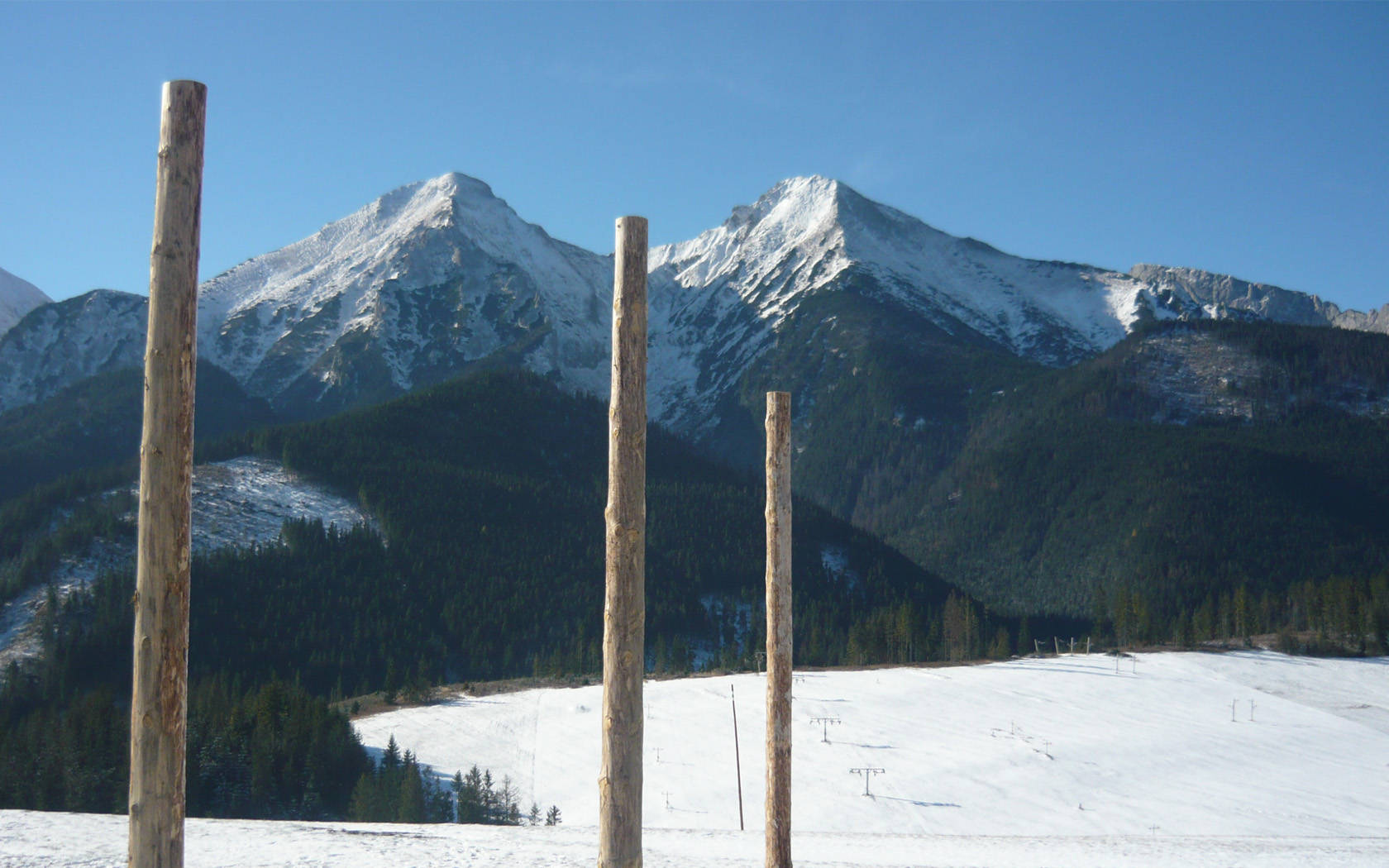 Slovakia 桌布 壁紙 — 三根風化的木柱框住了一片廣闊的雪山景觀，背景是清澈的藍天。