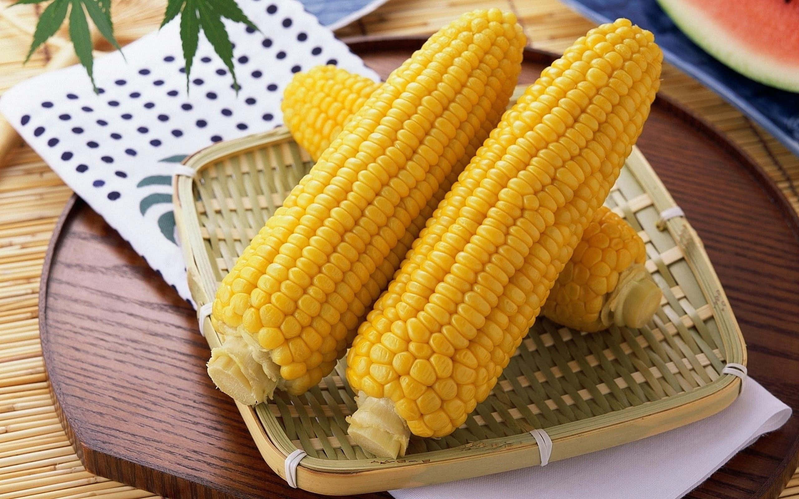Yellow Corn 桌布 壁紙 — 三根鮮黃色的玉米放在木托盤上的編織竹籃中，旁邊有一片西瓜和葉狀裝飾。