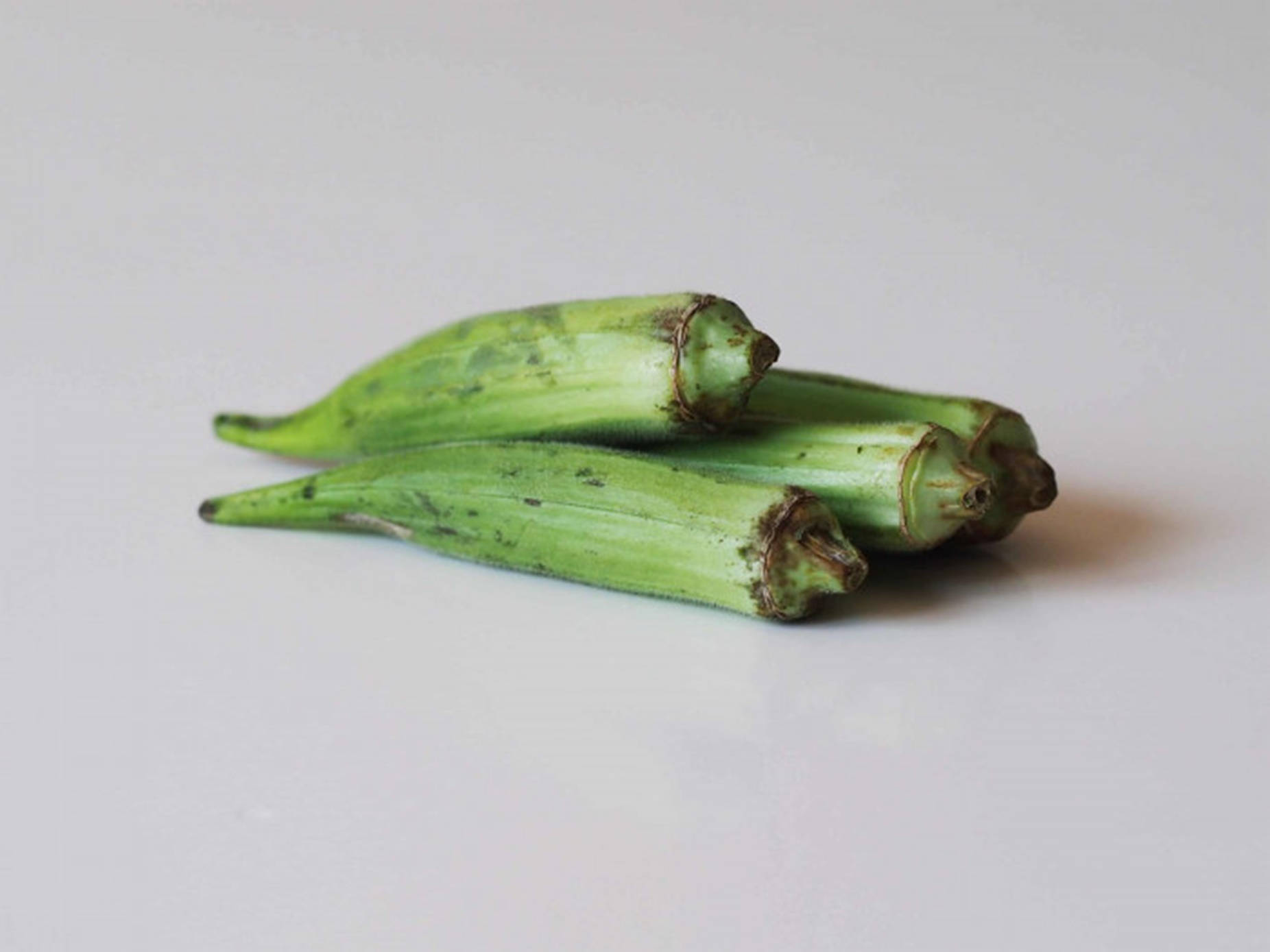 Vegetables 桌布 壁紙 — 三條新鮮的綠色秋葵莢簡單地堆放在明亮的白色表面上，帶有柔和陰影。