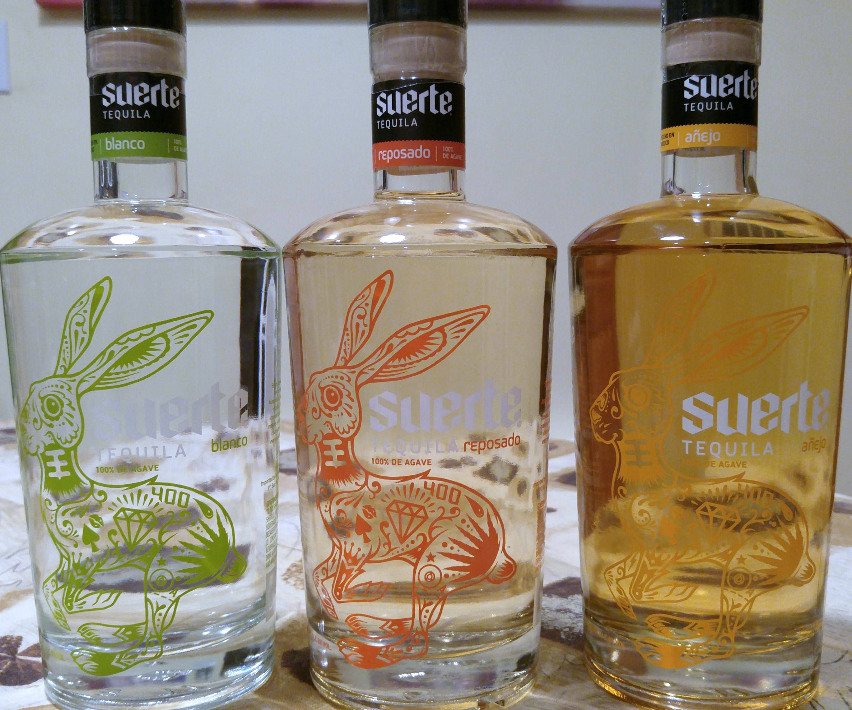 alcohol 桌布 壁紙 — 三款Suerte龍舌蘭酒瓶（blanco、reposado和añejo）並排展示，呈現綠色、橙色和金色色調中精緻的兔子設計。