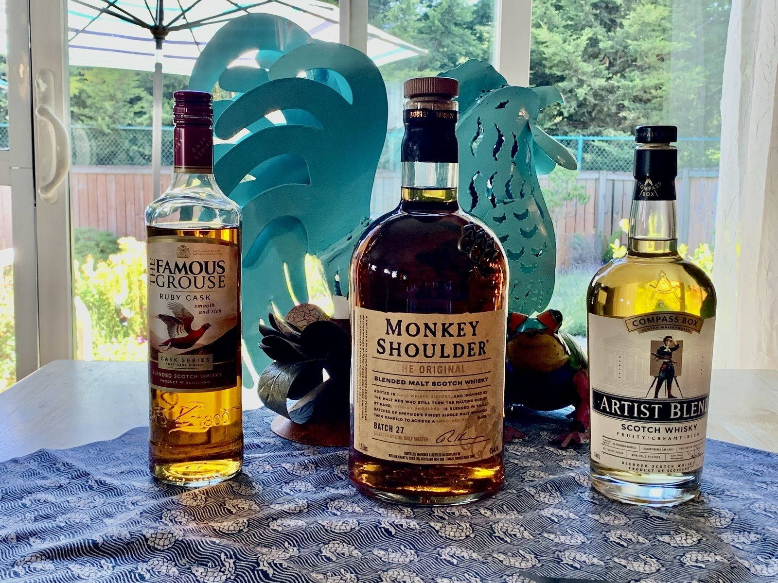 alcohol 桌布 壁紙 — 三款不同的蘇格蘭威士忌酒瓶放在窗邊的藍色圖案桌布上，窗外有綠植。