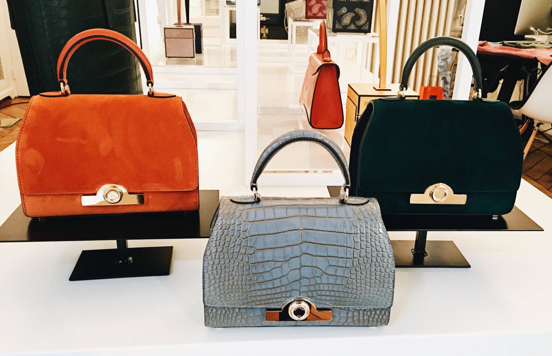 Luxury Brands 桌布 壁紙 — 三款奢華手袋，採用橙色、藍綠色和灰色鱷魚皮紋理，在明亮的精品店中展示於黑色支架上。