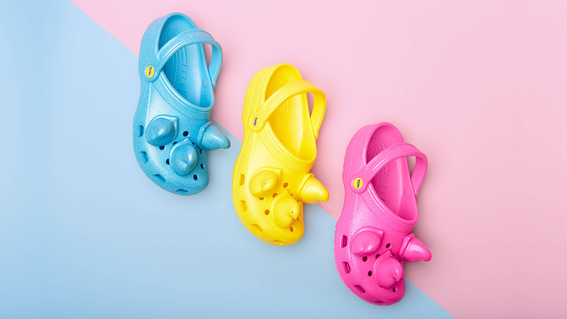 Crocs 桌布 壁紙 — 三款色彩繽紛的犀牛主題Crocs鞋，分別為藍色、黃色和粉紅色，以對角線排列在粉彩粉紅和藍色背景上。