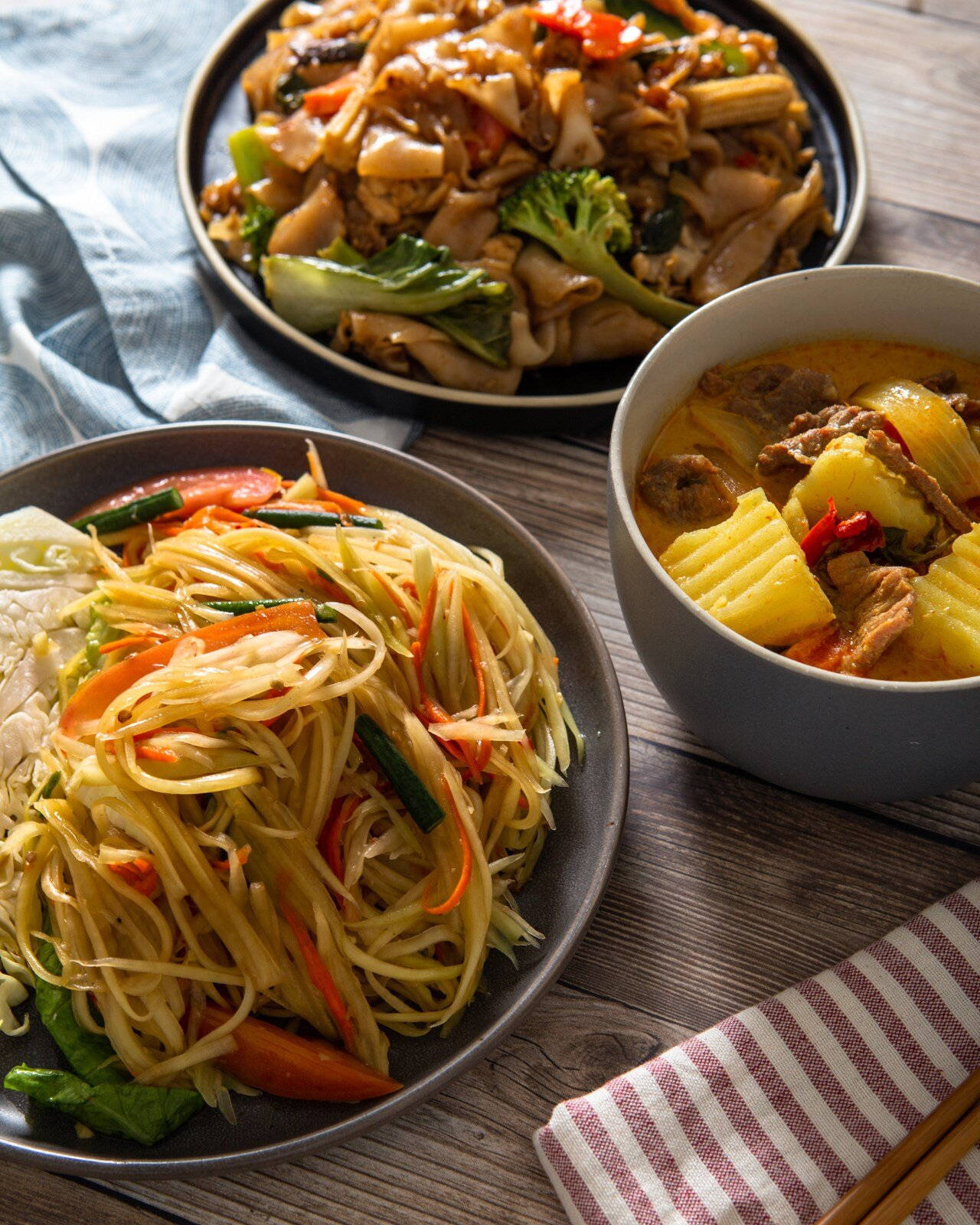 Healthy Food 桌布 壁紙 — 三款色彩鮮豔的亞洲菜餚，包括木瓜沙拉、咖哩和炒菜，擺放在質樸的木桌上。