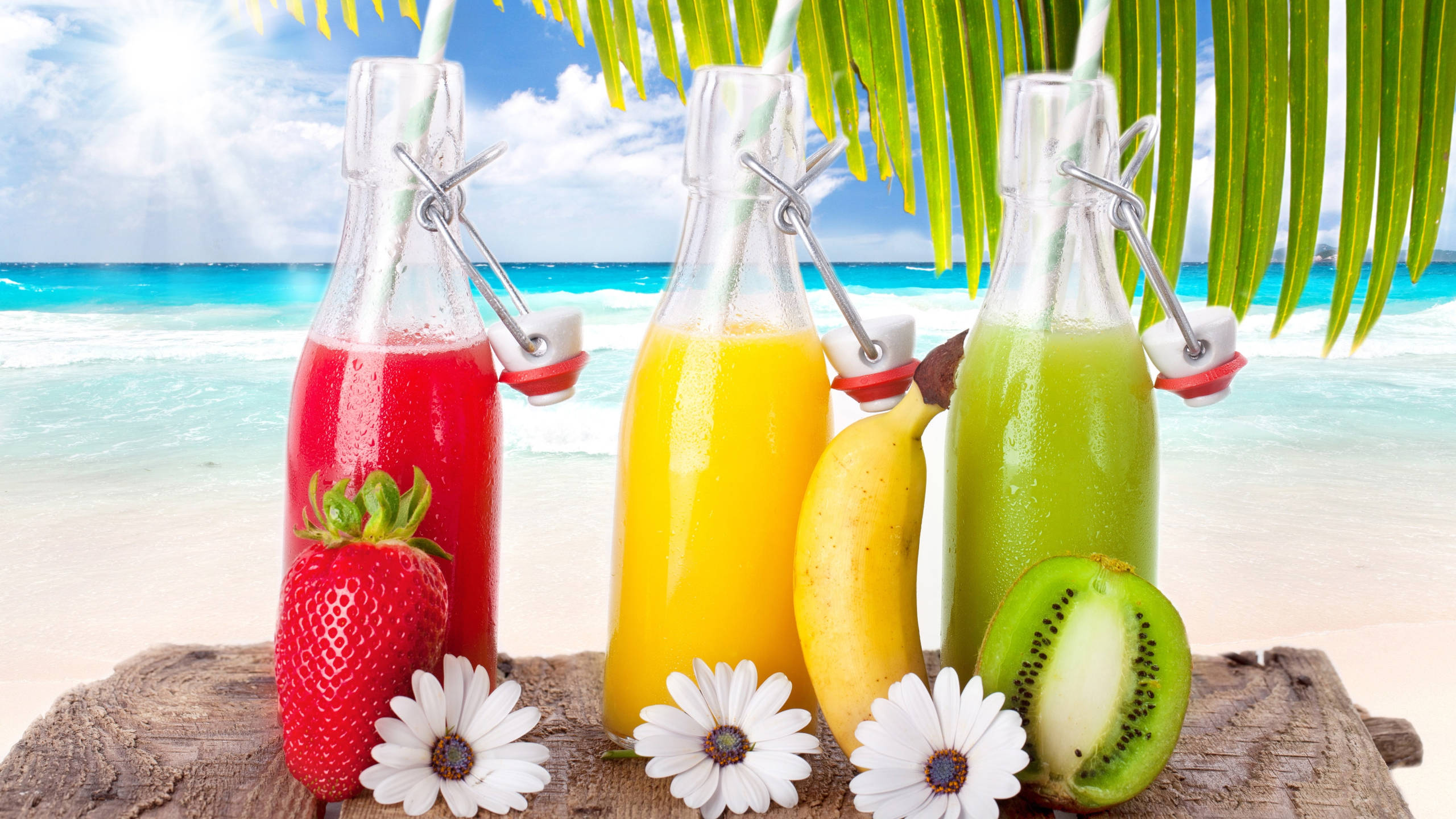 Tropical Drink 桌布 壁紙 — 三款色彩鮮豔的果汁裝在玻璃瓶中，放在木板上，旁邊是新鮮水果和鮮花，背景是陽光明媚的熱帶海灘。