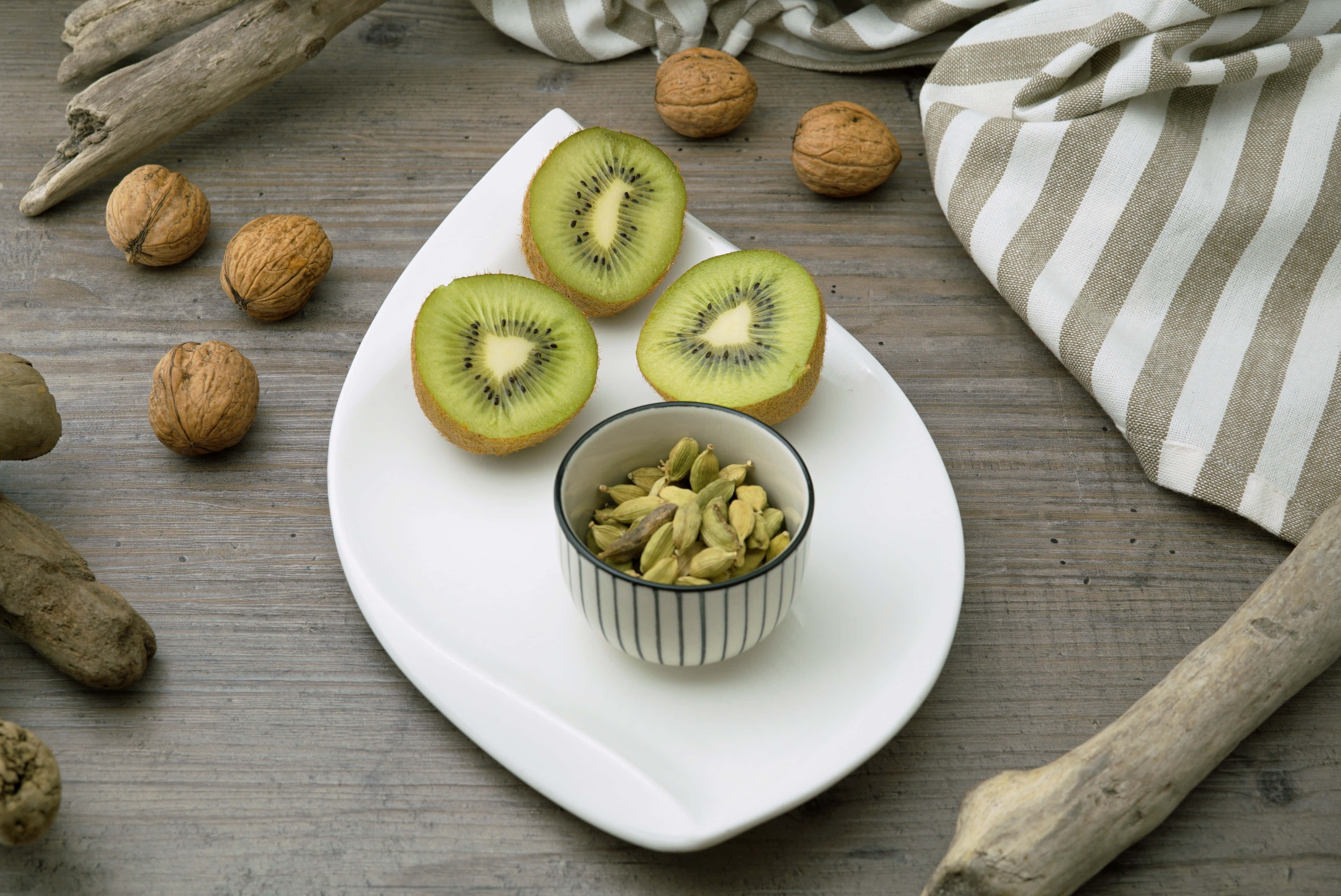 Kiwi 桌布 壁紙 — 三片奇異果和一碗小荳蔻籽放在白色盤子上，旁邊有核桃和浮木，置於風化的木質表面上。