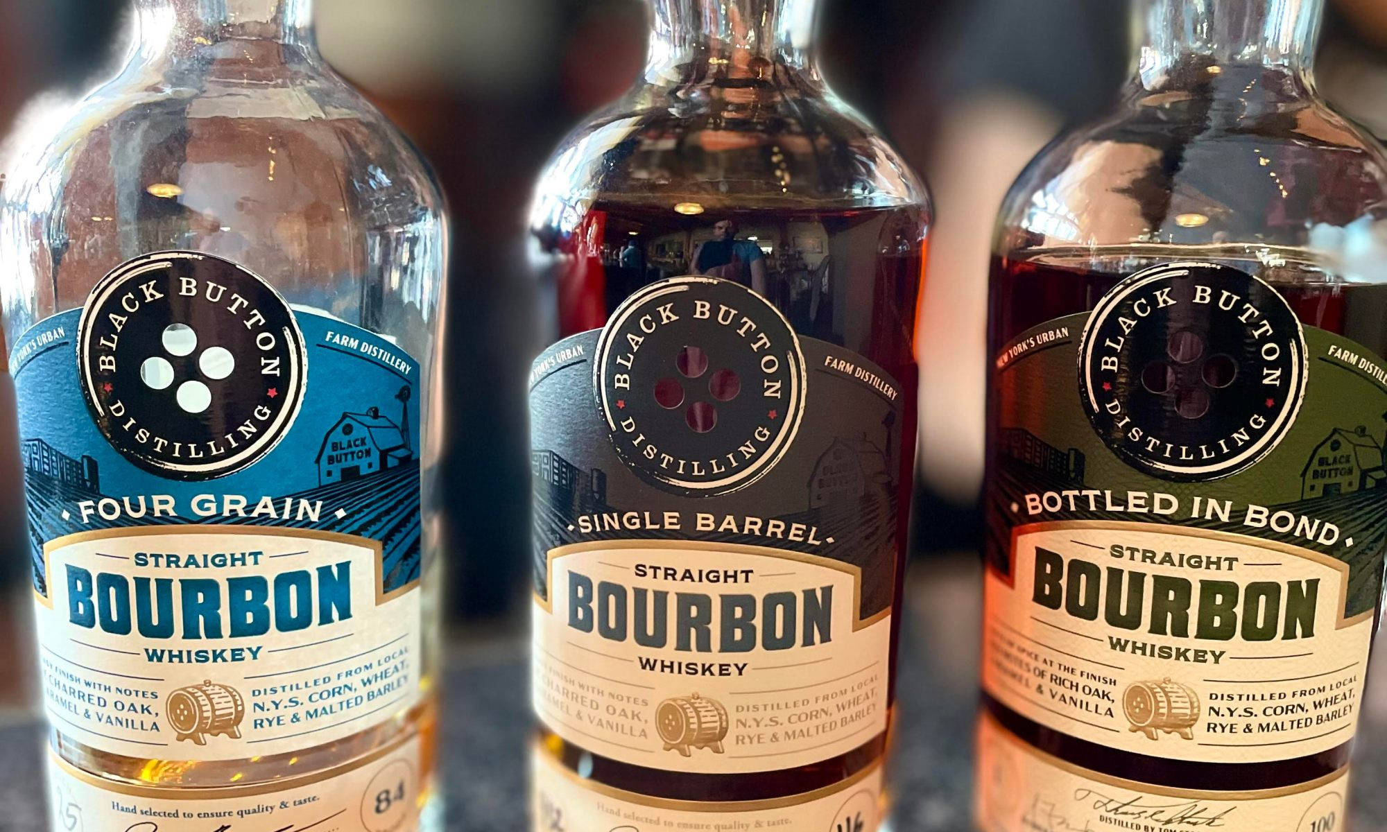 alcohol 桌布 壁紙 — 三瓶 Black Button Distilling 波本威士忌，帶有獨特的標籤和液體顏色，在深色表面上排成一排展示。