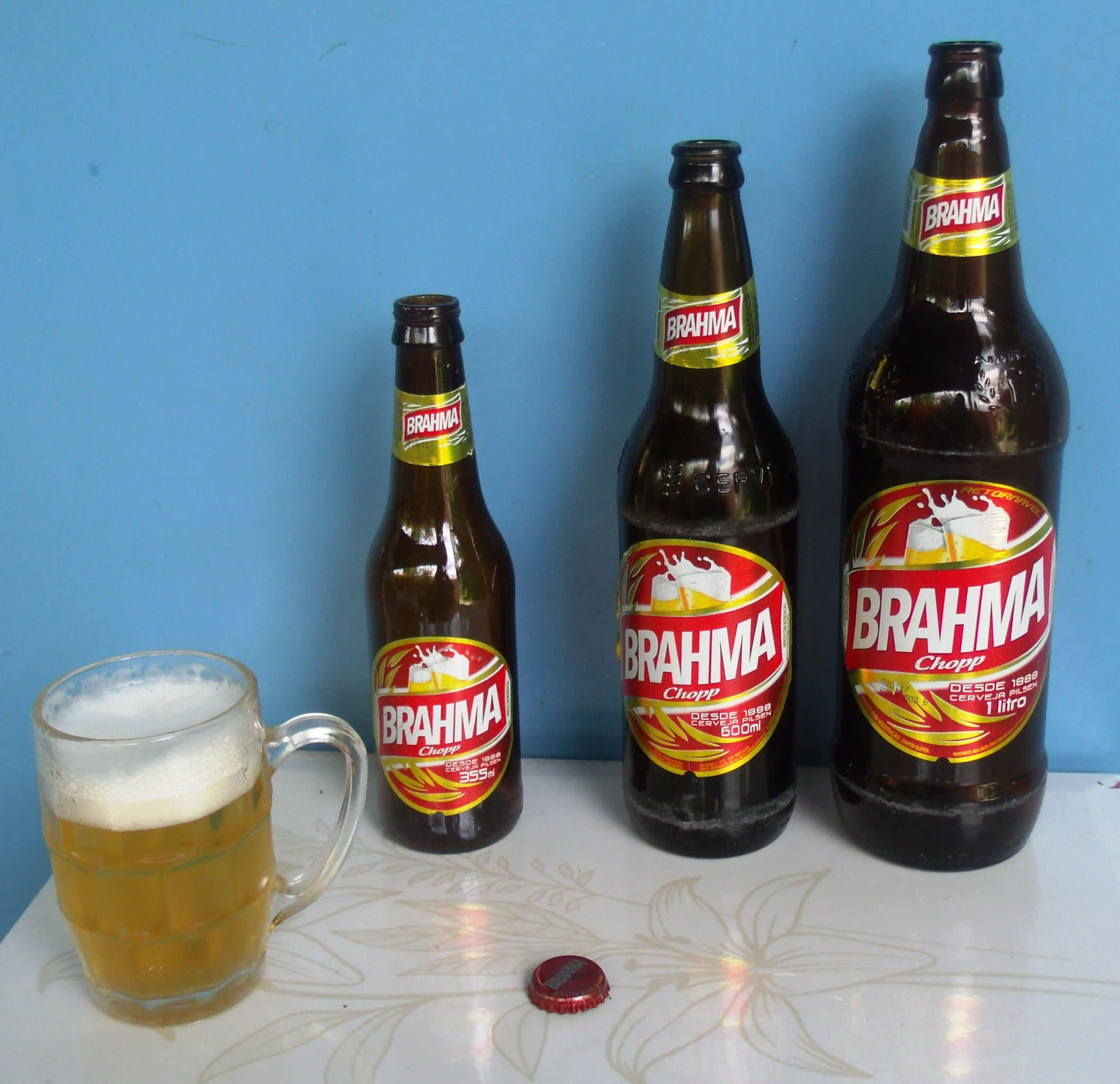 Drink 桌布 壁紙 — 三瓶 Brahma 啤酒和一個有泡的馬克杯放在花卉圖案的桌子上，背景是純藍色的牆壁。