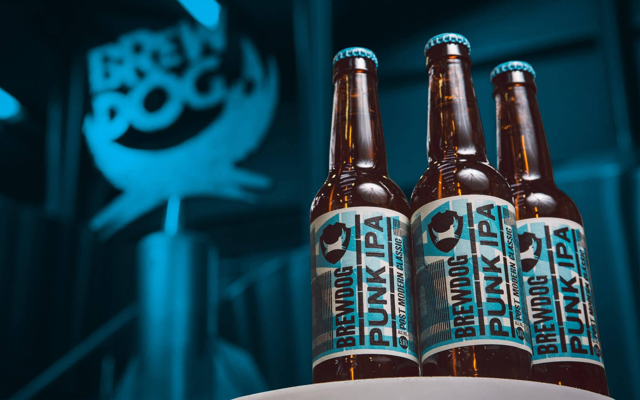 Drink 桌布 壁紙 — 三瓶 BrewDog Punk IPA 啤酒在發光的藍色 BrewDog 標誌背景前清晰對焦。
