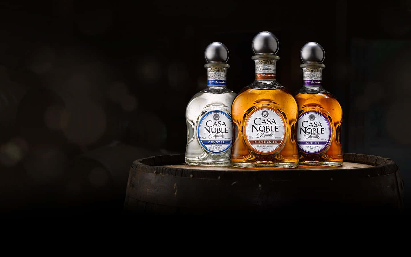 Tequila 桌布 壁紙 — 三瓶 Casa Noble 龍舌蘭酒，包括水晶 reposado 和 añejo 品種，優雅地放置在一個風化的木桶上，背景黑暗模糊。