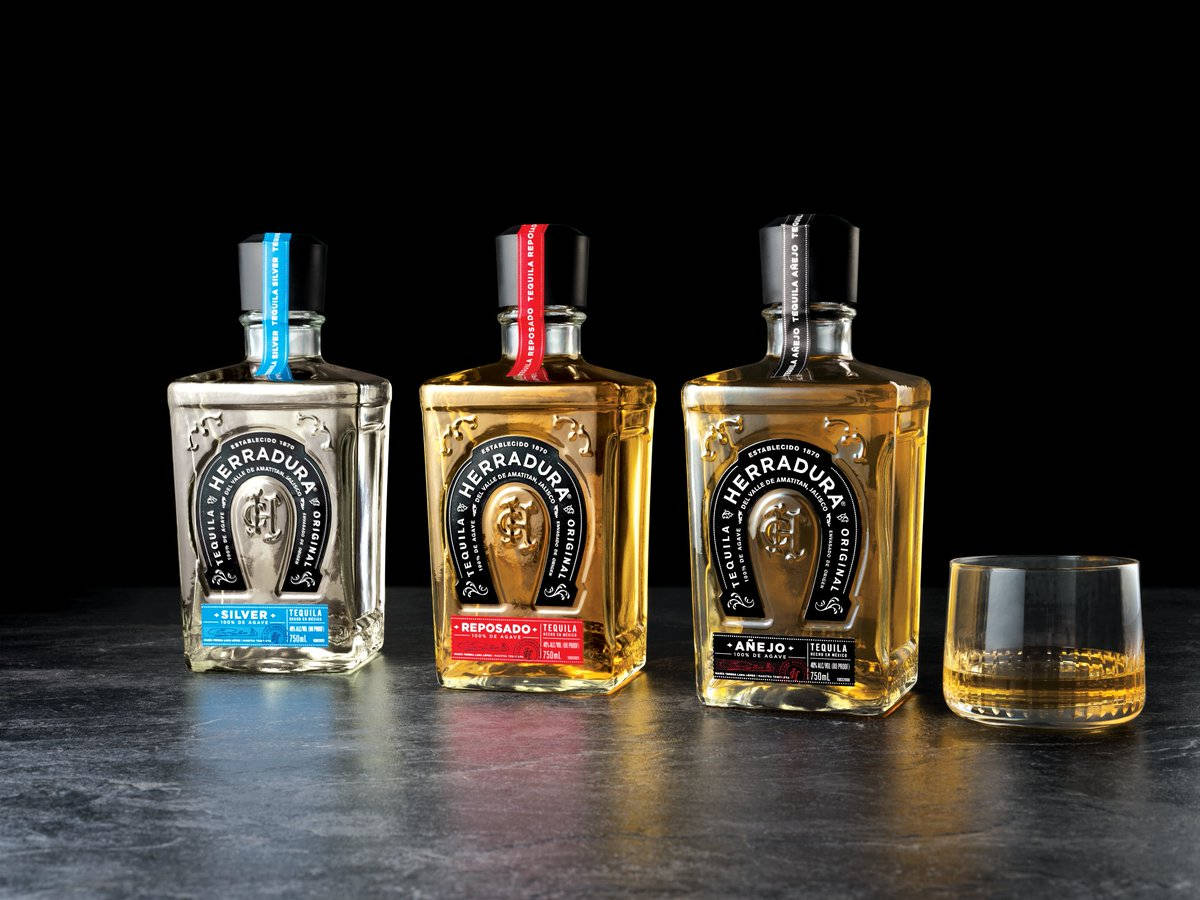 手機 桌布 壁紙 — 三瓶 Herradura 龍舌蘭酒和一杯琥珀色液體放在黑色背景的深色紋理表面上。