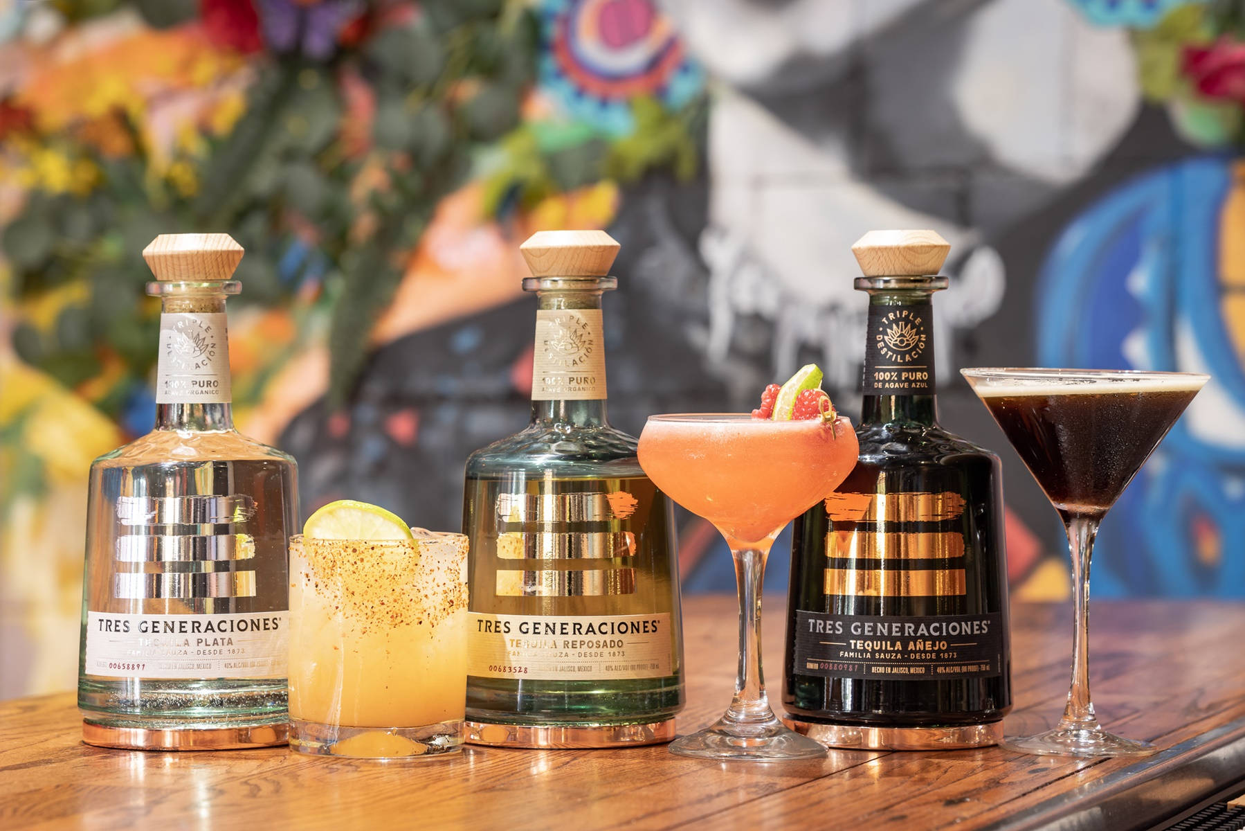 alcohol 桌布 壁紙 — 三瓶 Tres Generaciones 龍舌蘭酒和雞尾酒展示在木製吧檯上，背景是色彩繽紛的壁畫。