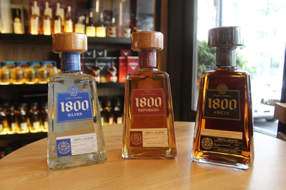 alcohol 桌布 壁紙 — 三瓶1800龍舌蘭酒（銀色、雷波薩多、阿涅霍）放在一個木製吧台上，後面有酒架。