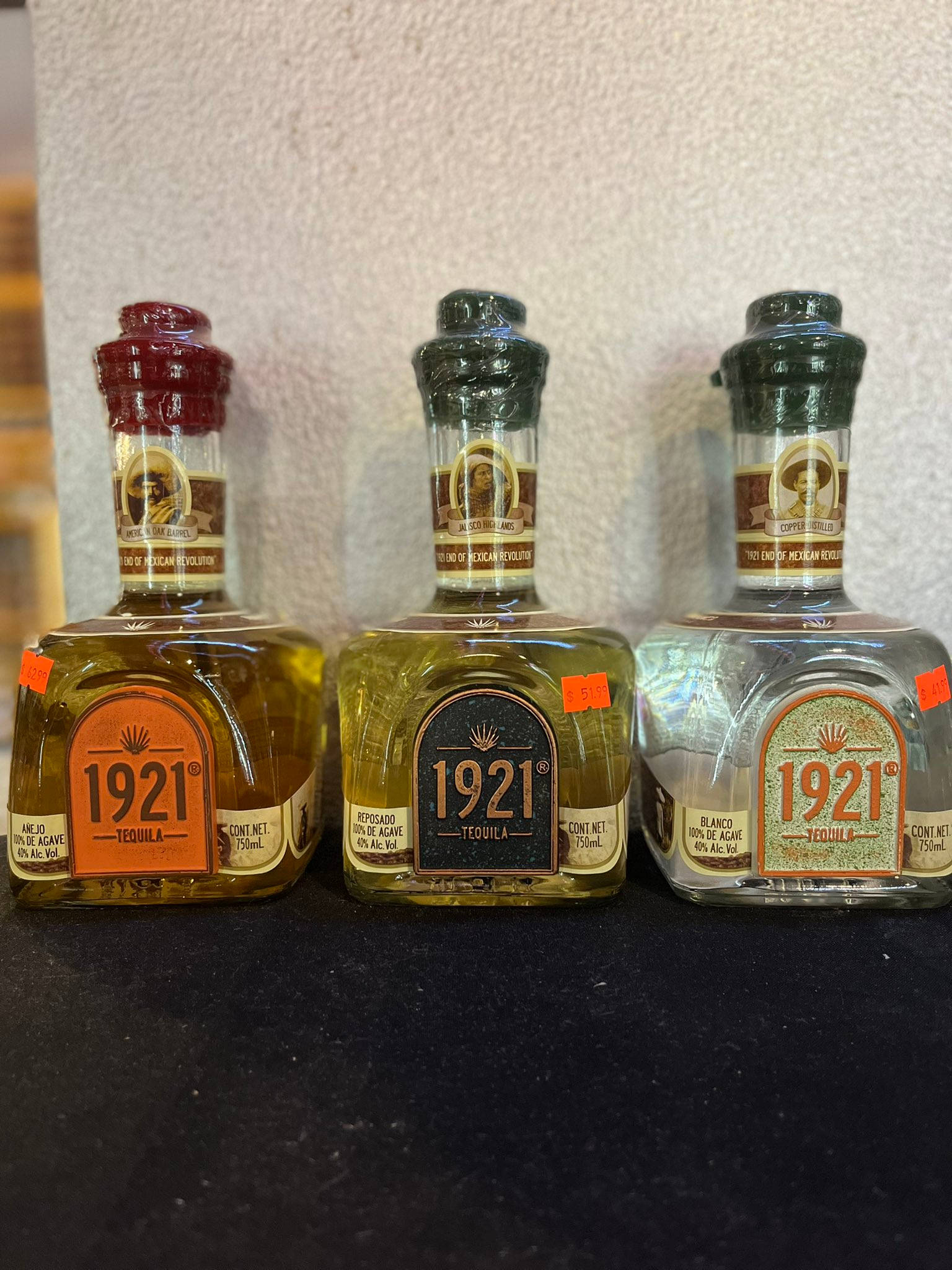 drinks 桌布 壁紙 — 三瓶1921年龍舌蘭酒，琥珀金銀玻璃瓶身，排列在深色表面上，背景為紋理牆壁。
