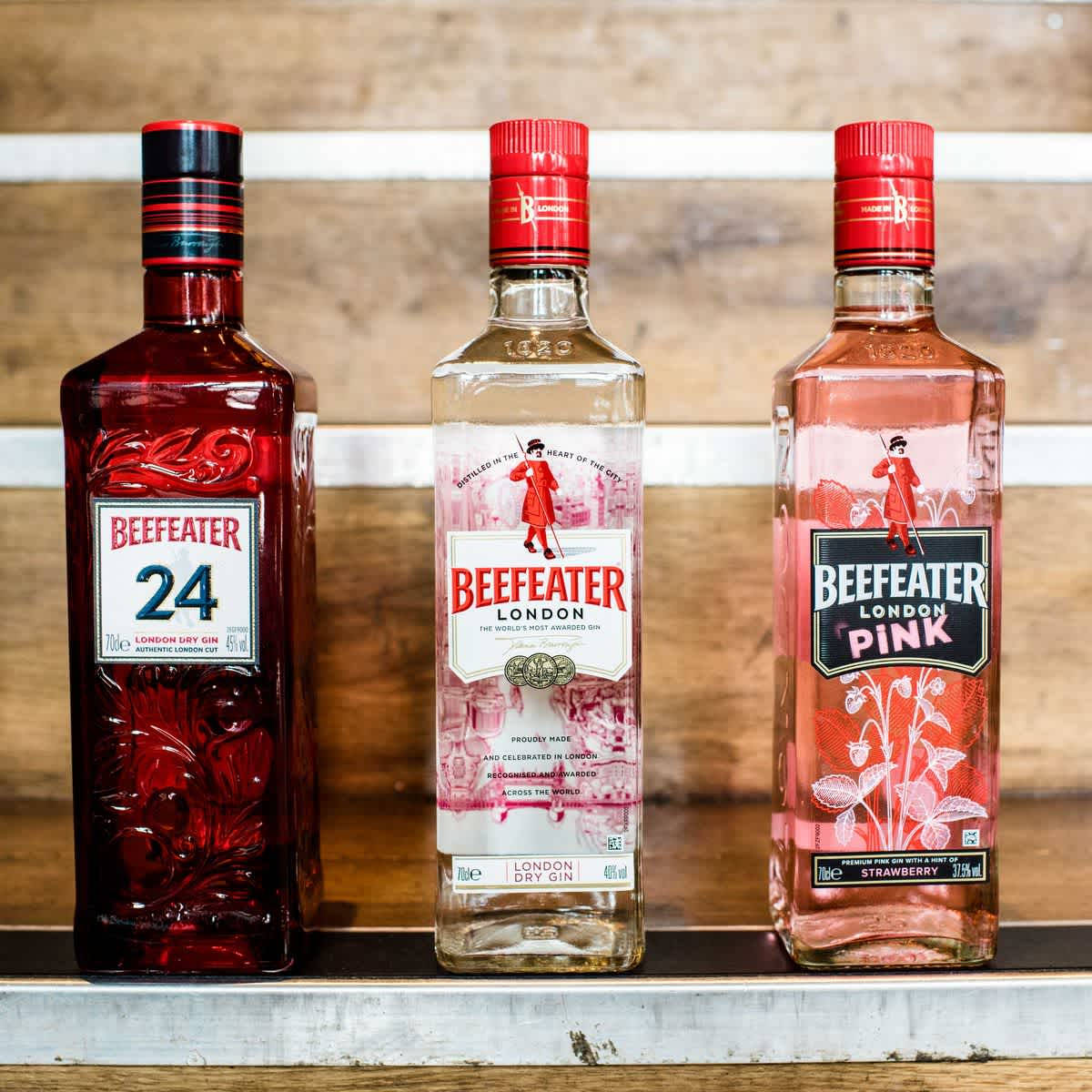 bottle 桌布 壁紙 — 三瓶Beefeater琴酒，分別呈現紅色、透明和粉紅色調，整齊地放在金屬架上，背景是質樸的木頭。