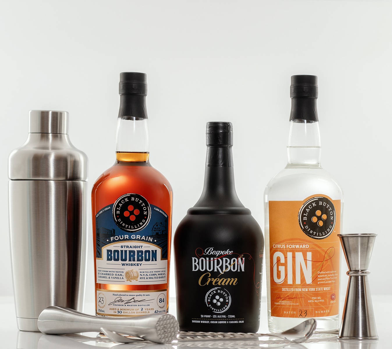 alcohol 桌布 壁紙 — 三瓶Black Button Distilling烈酒和酒吧工具整齊地排列在白色表面上，背景乾淨。