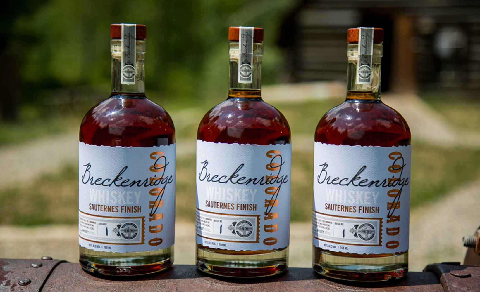 drinks 桌布 壁紙 — 三瓶Breckenridge Whiskey Sauternes Finish並排放在生鏽的金屬表面上，背景是模糊的綠色戶外景色。