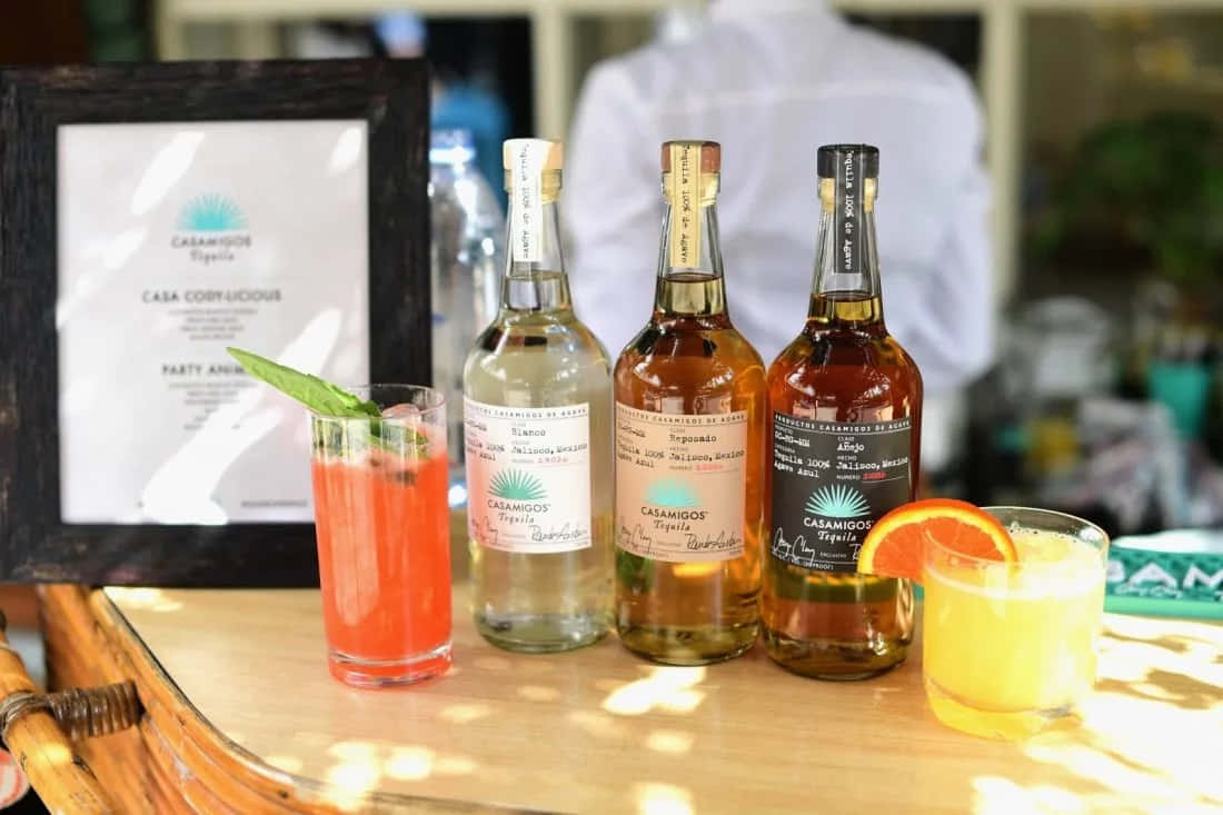 alcohol 桌布 壁紙 — 三瓶Casa Cinco龍舌蘭酒和兩杯色彩繽紛的雞尾酒放在木製吧檯上，陽光普照。