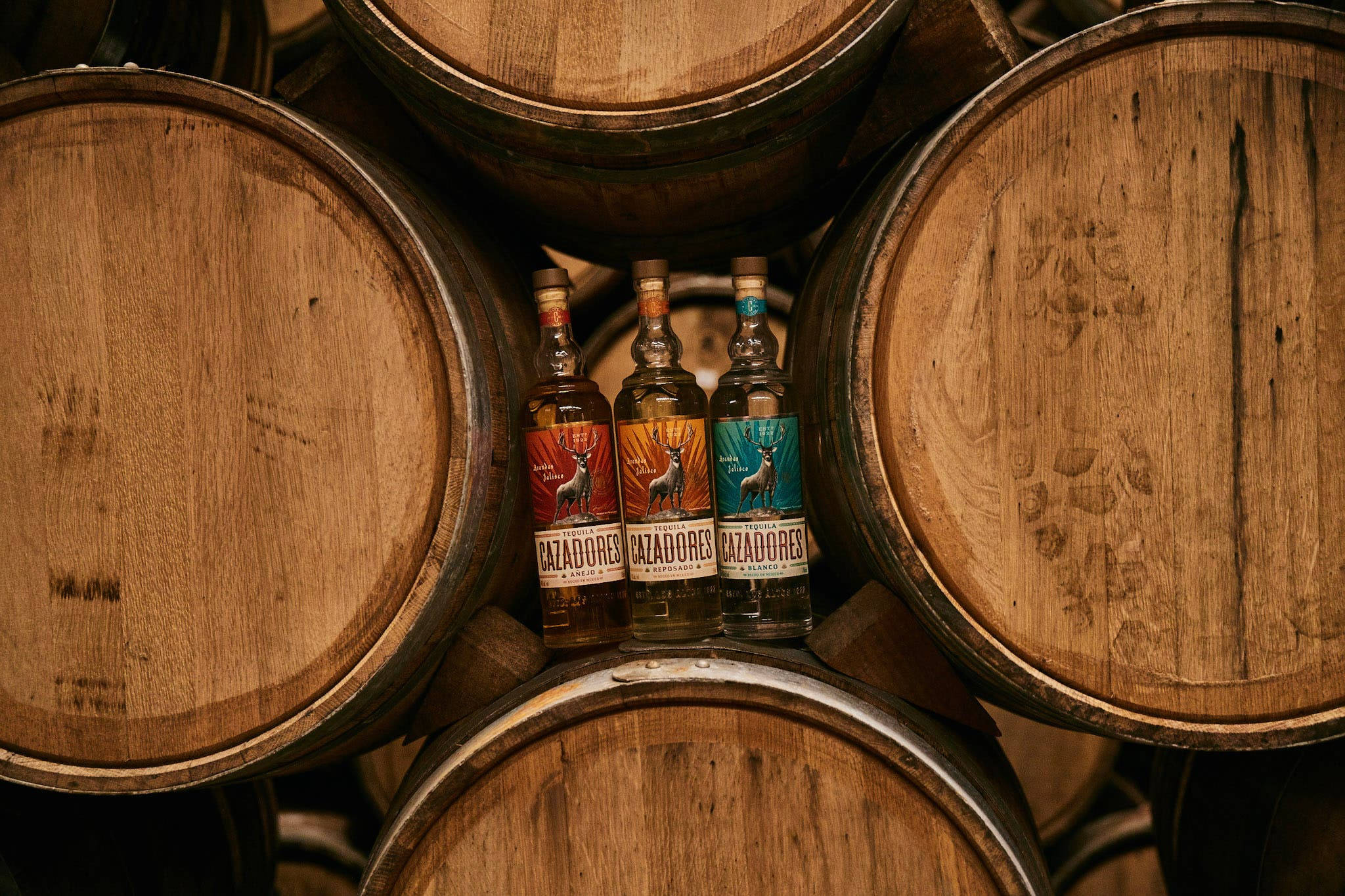 飲料 桌布 壁紙 — 三瓶Cazadores龍舌蘭酒放置在昏暗酒廠中堆疊的風化木桶之間。