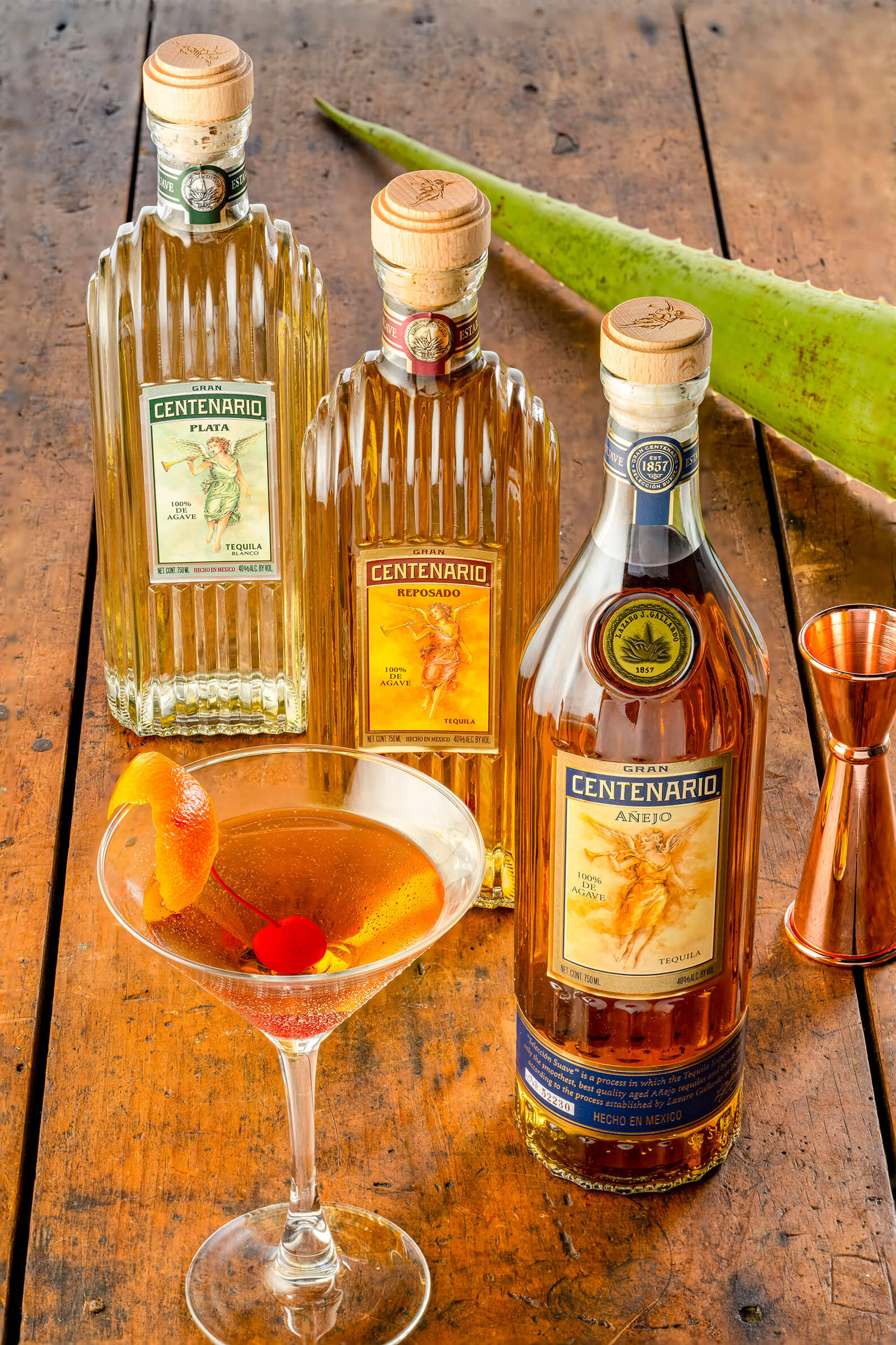 cocktail 桌布 壁紙 — 三瓶Centenario龍舌蘭酒和一杯以柳橙和櫻桃裝飾的雞尾酒，放在一張質樸的木桌上，旁邊有一片蘆薈葉。