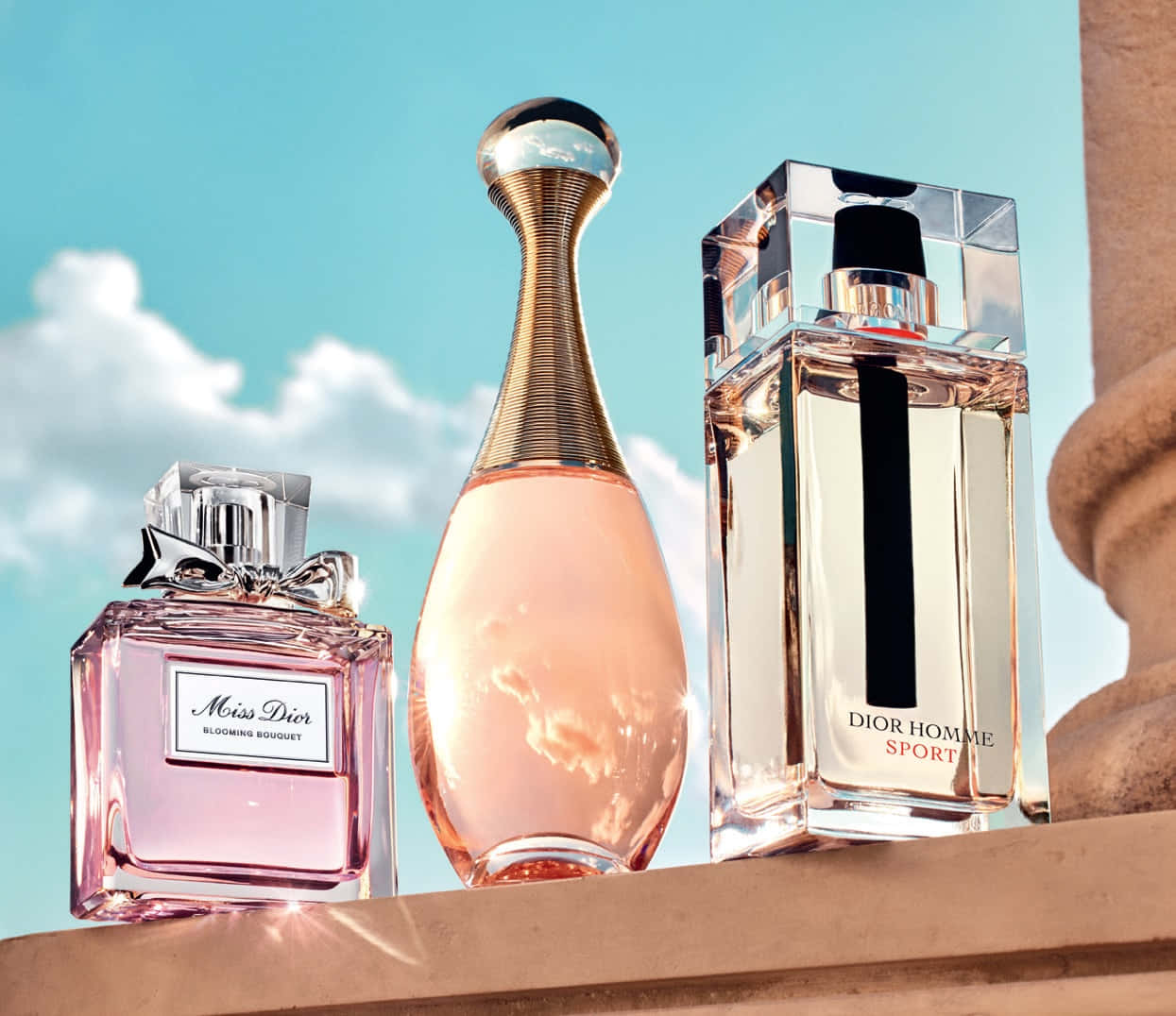 Fragrance 桌布 壁紙 — 三瓶Dior香水在明亮的陽光下閃閃發光，背景是晴朗的藍天和蓬鬆的白雲。