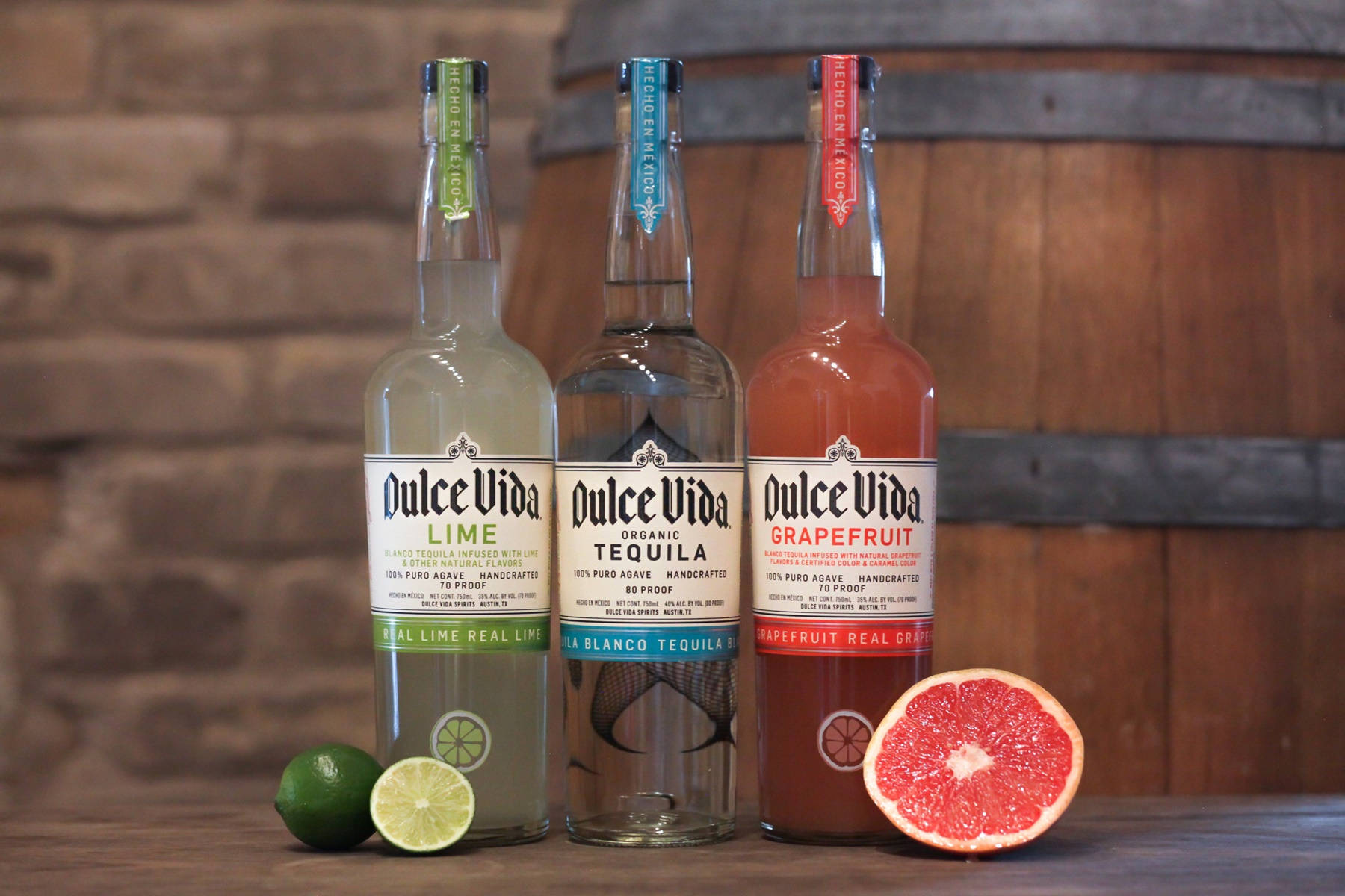 cocktail 桌布 壁紙 — 三瓶Dulce Vida龍舌蘭酒（萊姆、葡萄柚及原味）放在木桌上，旁邊有新鮮柑橘類水果，背景是質樸的磚牆和木桶。
