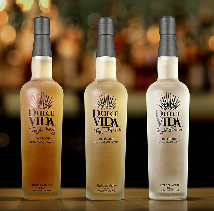 cocktail 桌布 壁紙 — 三瓶Dulce Vida龍舌蘭酒，呈現金色、琥珀色、透明和白色色調，整齊排列在木製吧檯上，背後有溫暖的散景燈光。