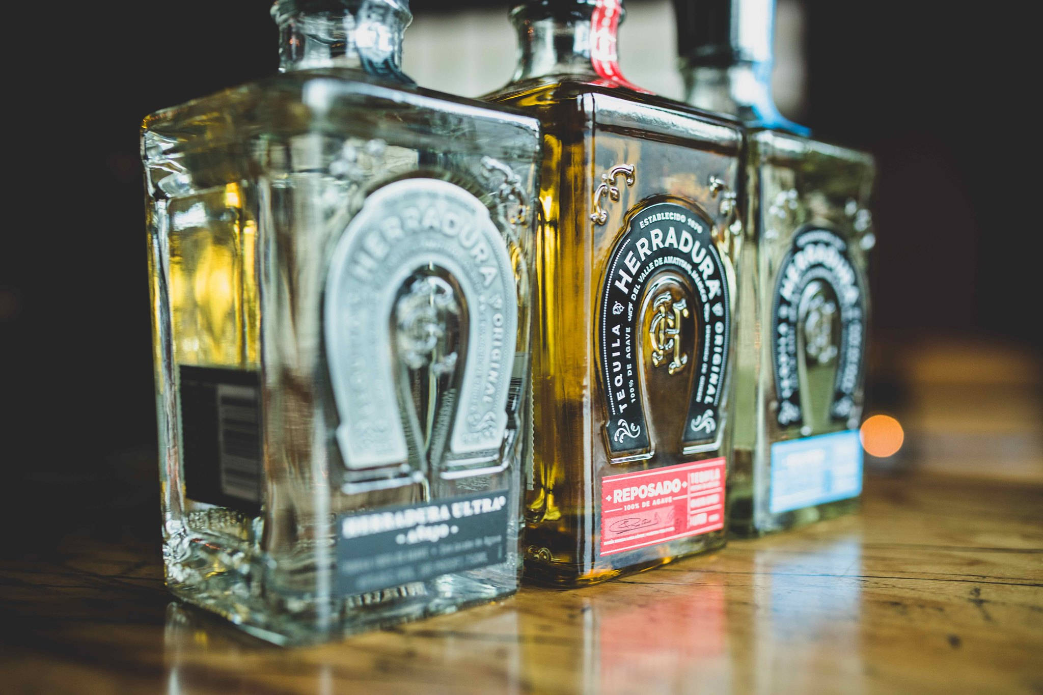 Herradura 桌布 壁紙 — 三瓶Herradura龍舌蘭酒整齊排列在木質吧檯上，背景是溫暖的散景燈光。