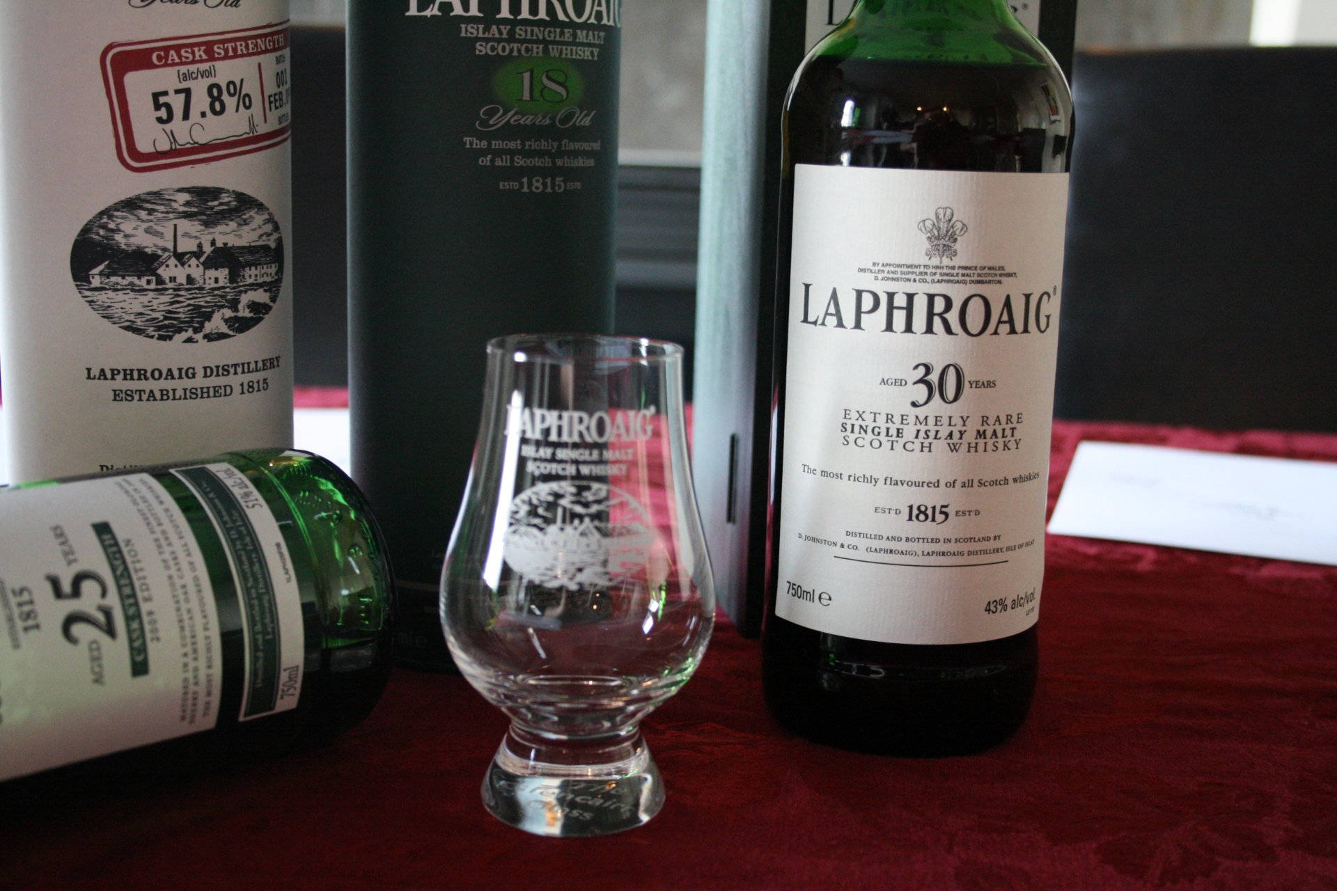 Wine 桌布 壁紙 — 三瓶Laphroaig威士忌和一個品酒杯擺放在深紅色天鵝絨表面上。