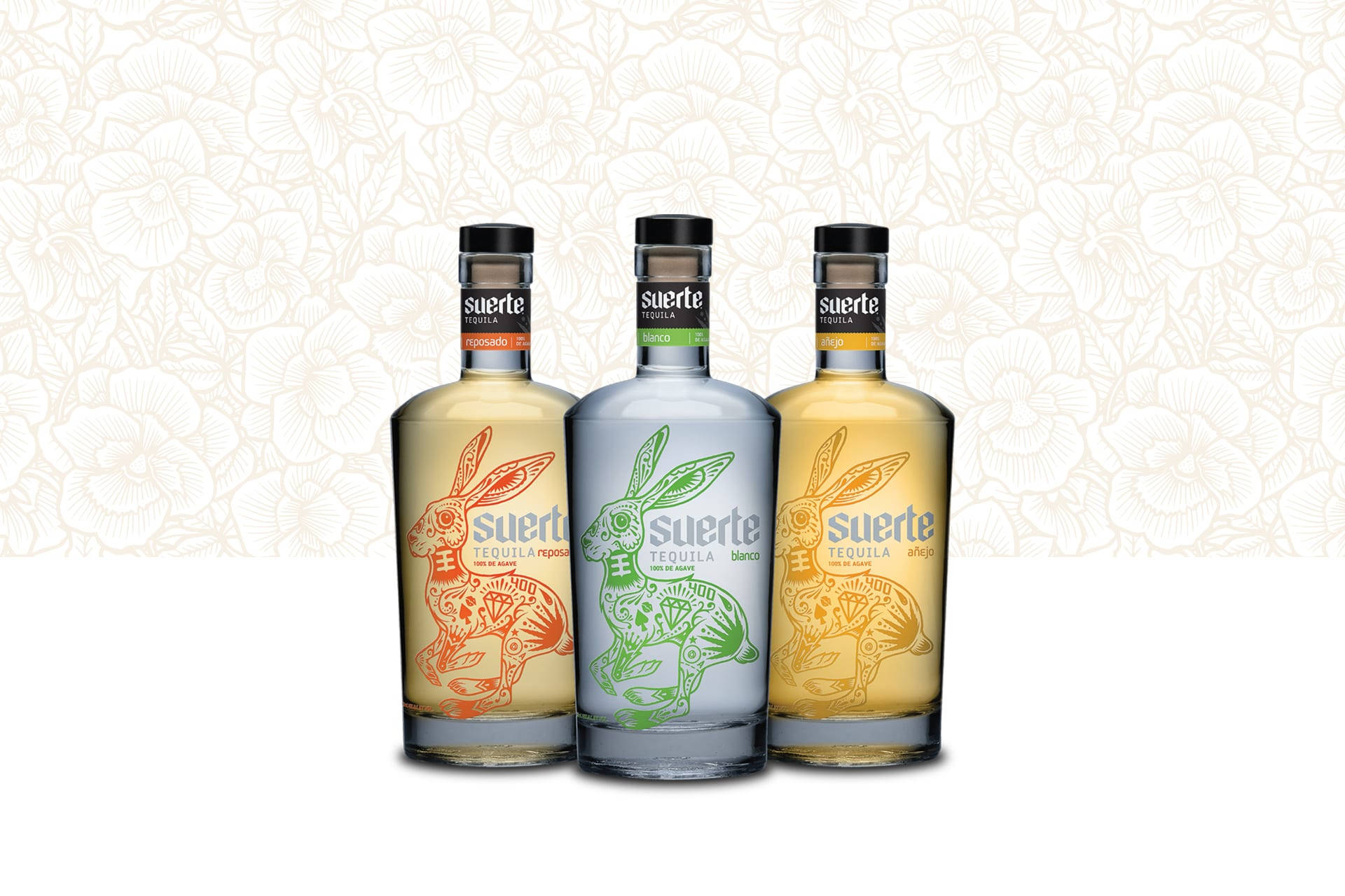 alcohol 桌布 壁紙 — 三瓶帶有兔子設計的Suerte龍舌蘭酒瓶，立在精緻的花卉壁紙背景前。