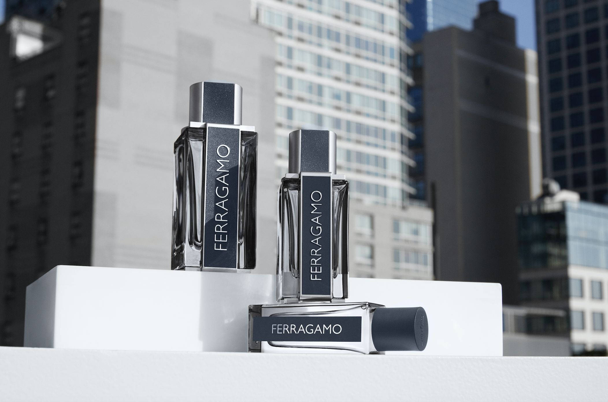 Ferragamo 桌布 壁紙 — 三瓶時尚的Ferragamo香水瓶放在白色方塊上，背景是明亮的日光下的現代城市摩天大樓。