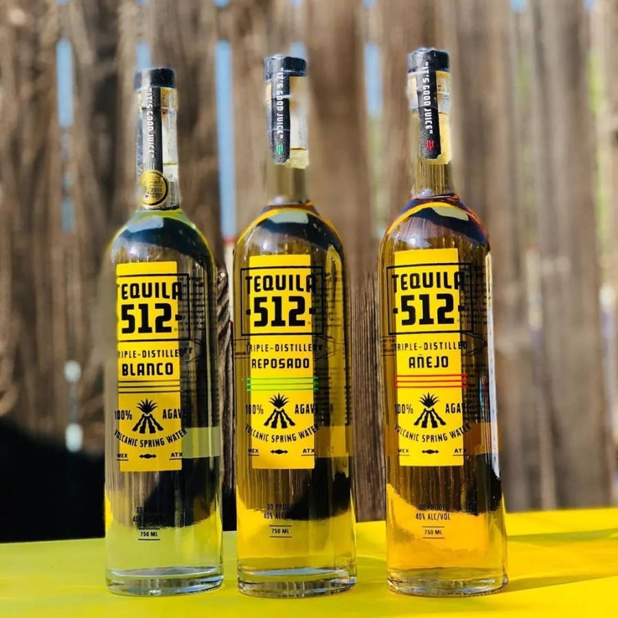 drinks 桌布 壁紙 — 三瓶標有 Blanco Reposado 和 Añejo 標籤的龍舌蘭酒瓶，在木柵欄背景前整齊排列在亮黃色的桌子上。