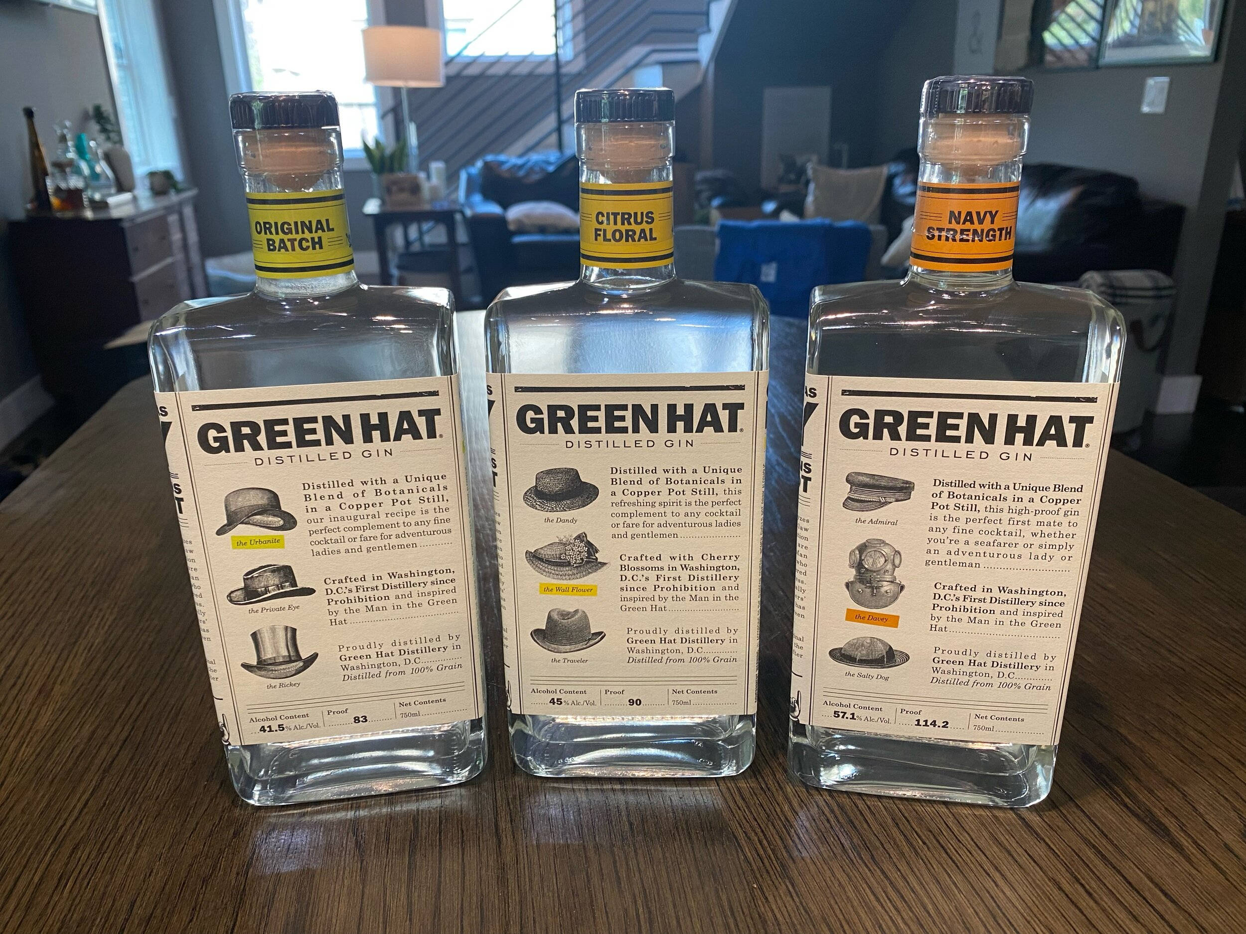alcohol 桌布 壁紙 — 三瓶透明的 Green Hat 蒸餾琴酒，帶有不同風味標籤，放在現代室內設計的木桌上。