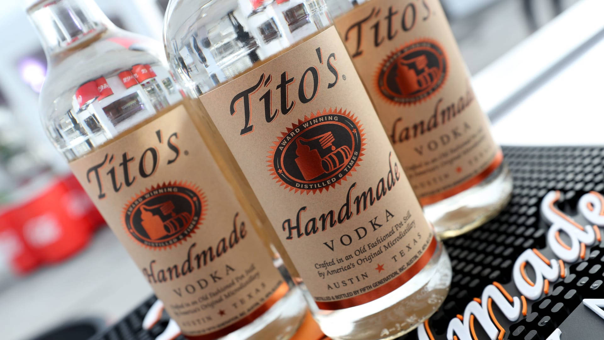 品牌 桌布 壁紙 — 三瓶透明的Tito's Handmade Vodka酒瓶傾斜放在黑色紋理的吧檯墊上，背景有一個模糊的紅色物體。