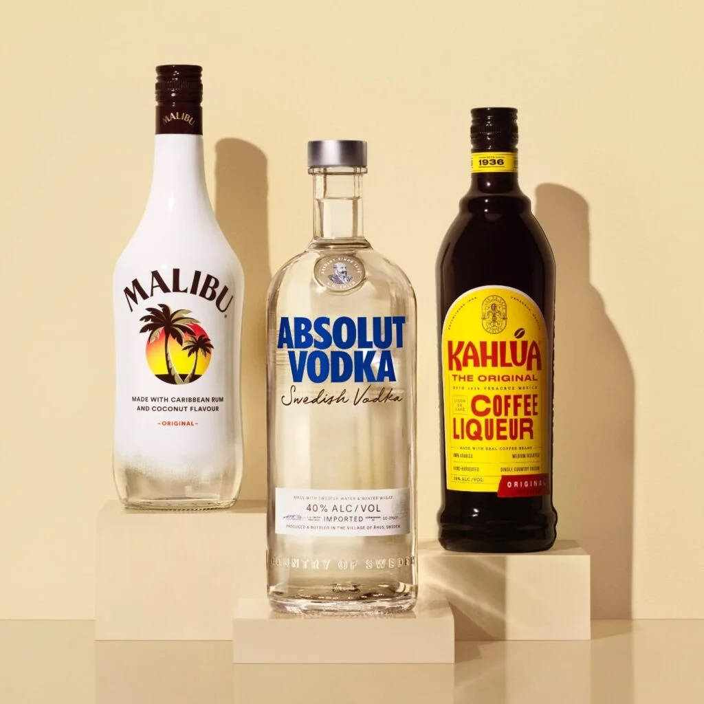 drinks 桌布 壁紙 — 三瓶雞尾酒 Malibu Absolut Vodka 和 Kahlua 優雅地展示在奶油色底座上，背景是溫暖的米色牆壁。