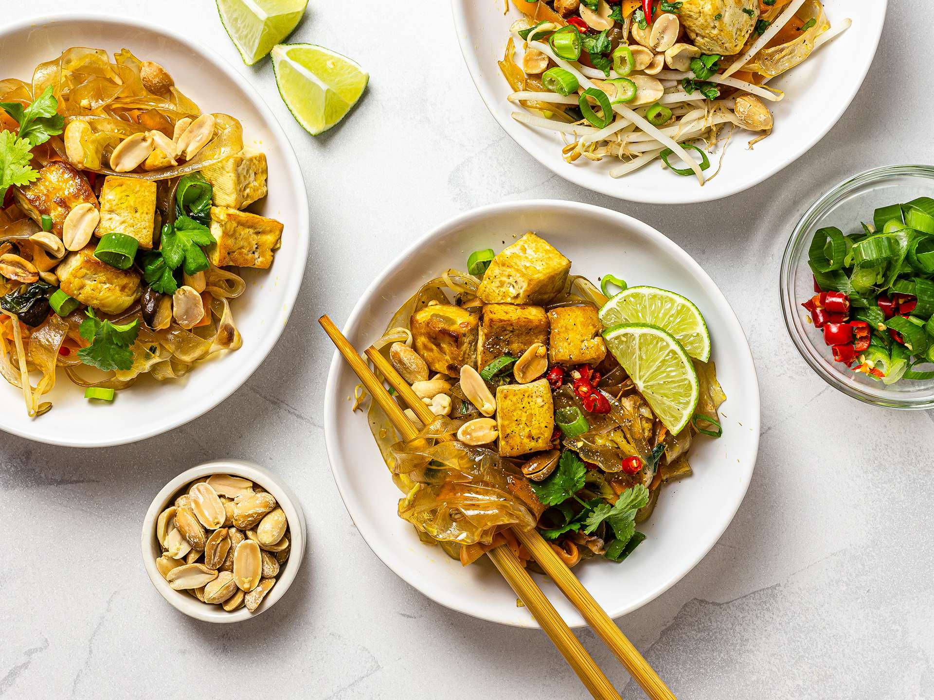 tofu 桌布 壁紙 — 三碗充滿活力的泰式炒米粉，上面放有豆腐、花生、萊姆和新鮮香草，置於淺灰色表面上。