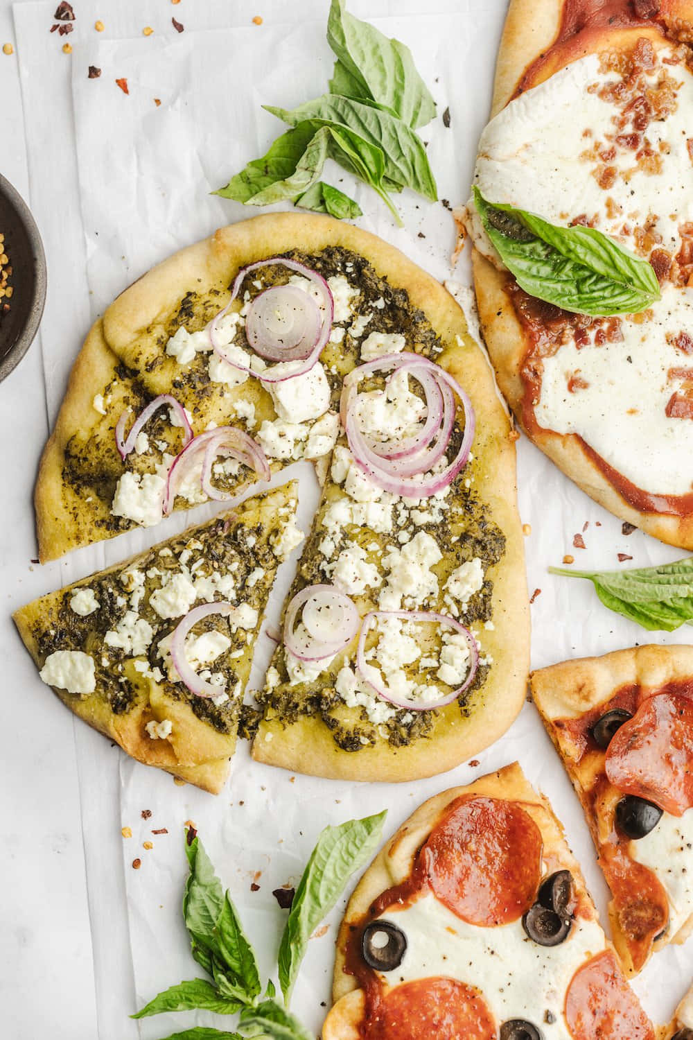 Pizza 桌布 壁紙 — 三種不同扁麵包披薩的俯拍，上面有香蒜醬、菲達起司、紅洋蔥、番茄、莫札瑞拉起司、義式辣香腸和橄欖，用新鮮羅勒裝飾，放在烘焙紙上。